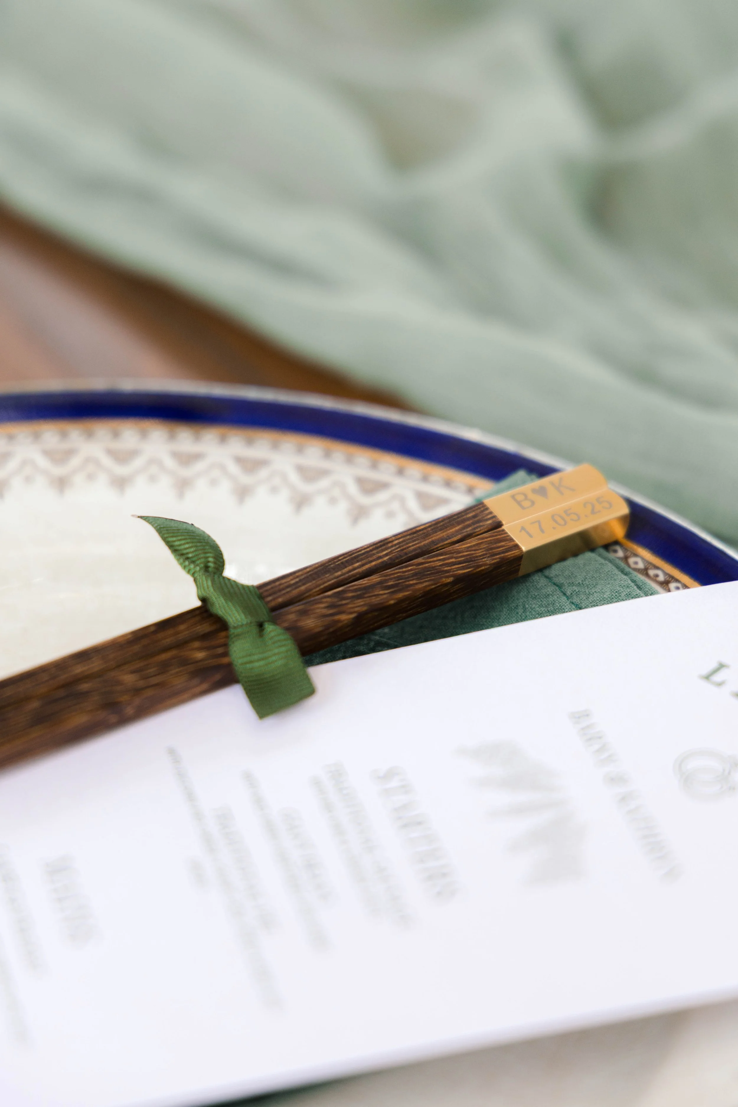 A close-up of a decorative wooden chopstick with a green ribbon tied around it, resting on a white paper menu or program. The chopstick has a beige band with handwritten initials and a date. In the background, there is a blue and gold edged plate and