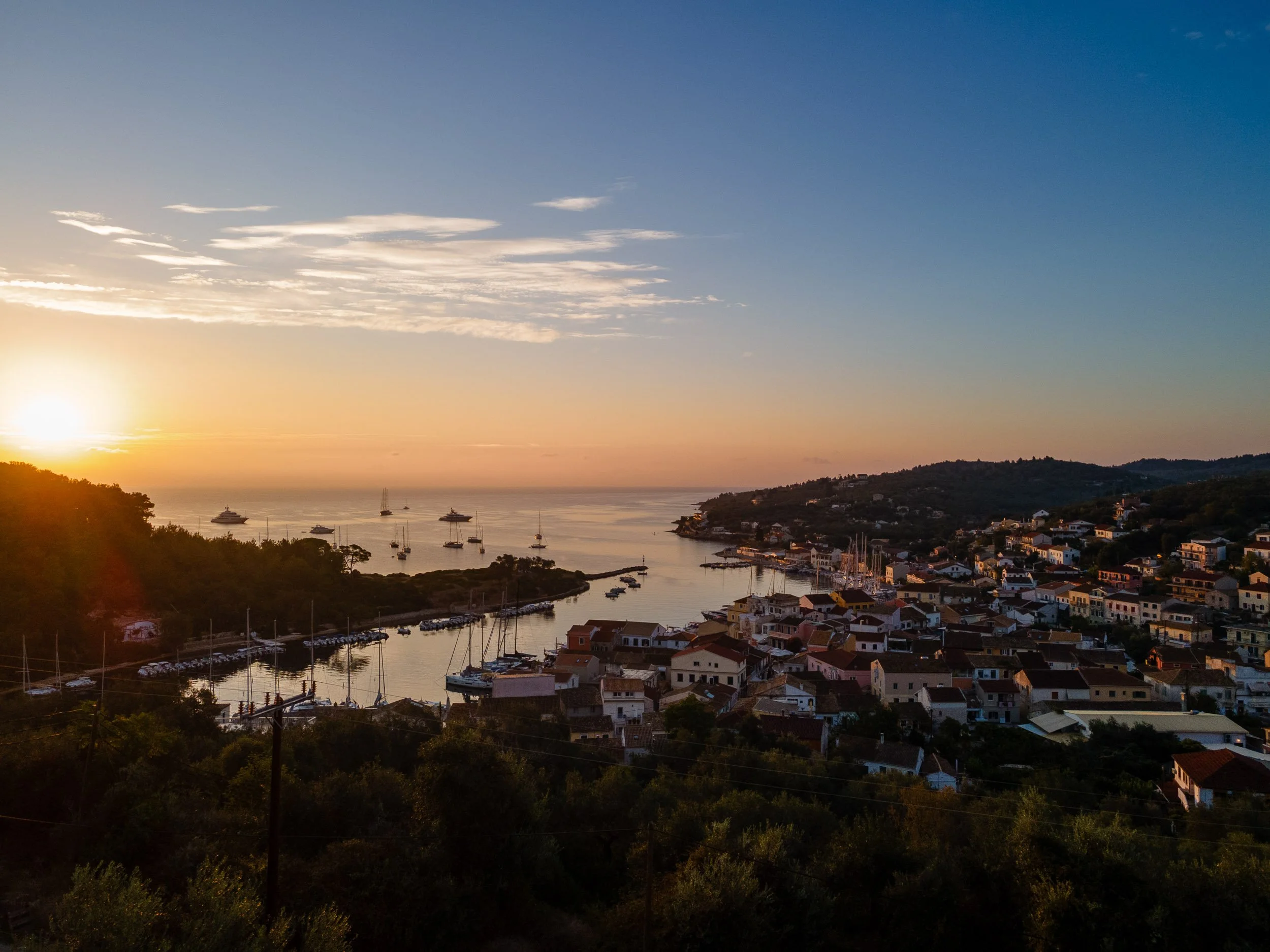 A scenic view of a harbor at sunset, with boats docked along the shoreline, houses on the hillside, and the sky with early evening colors.