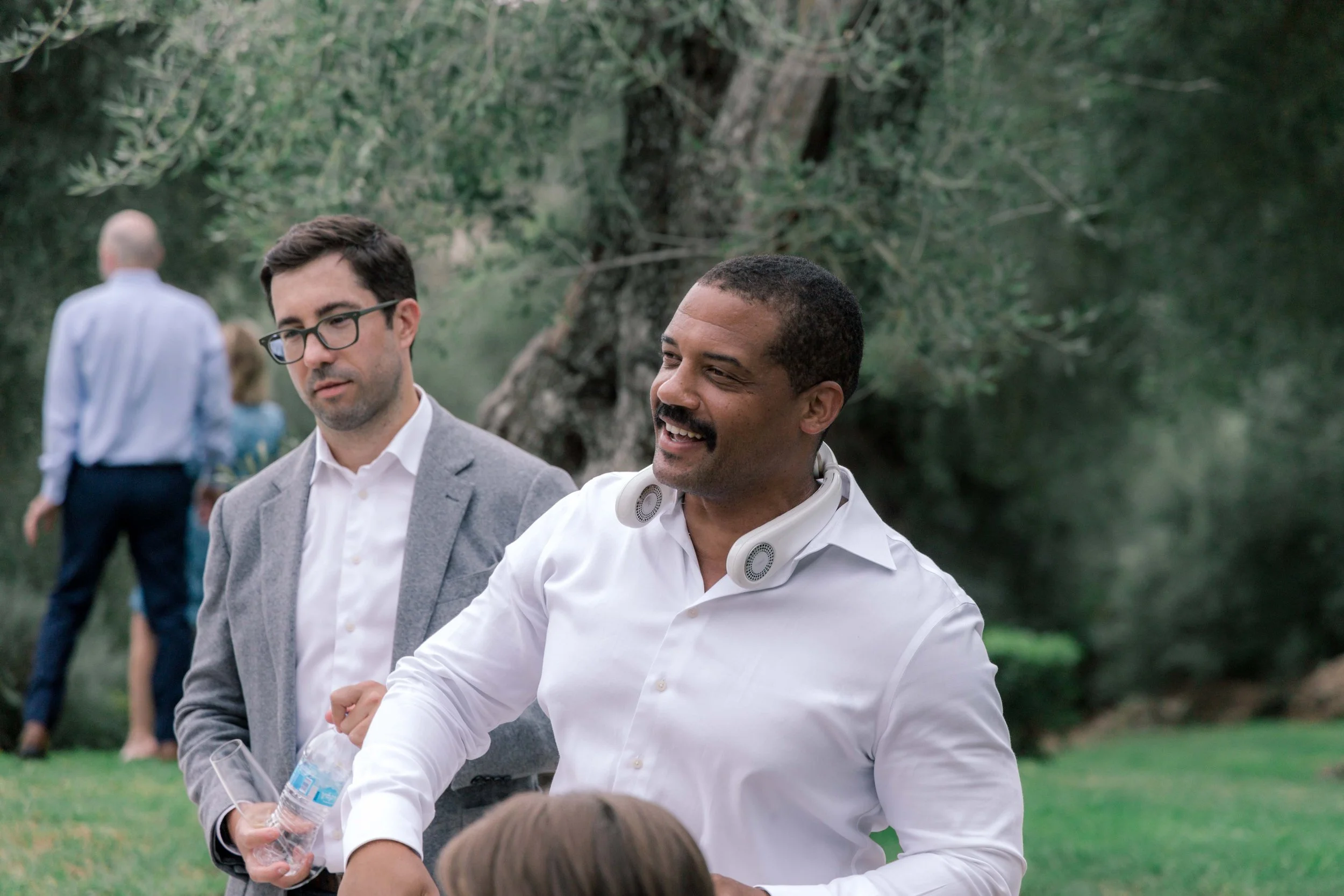 Two men, one in a gray suit and one in a white shirt with a headset around his neck, standing outdoors near trees, engaging in conversation.