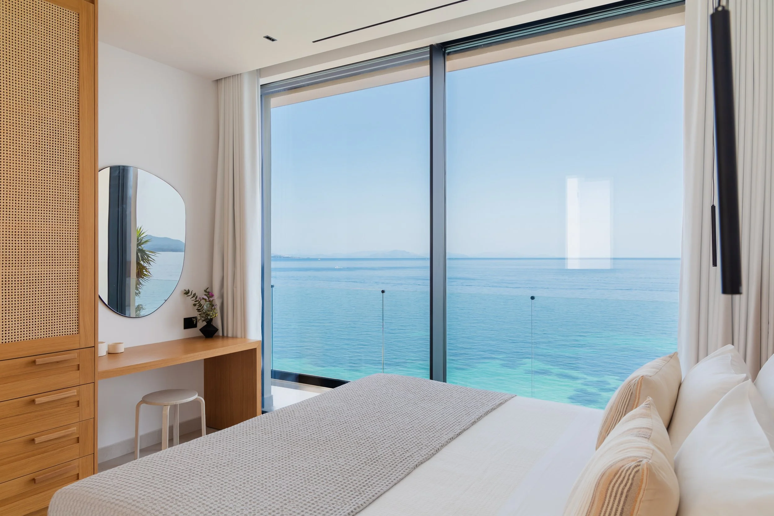 A modern bedroom with a large glass window offering a view of the ocean and distant mountains, a white bed with beige pillows, a wooden desk with a small stool, a mirror, and a small plant, with white curtains on either side of the window.