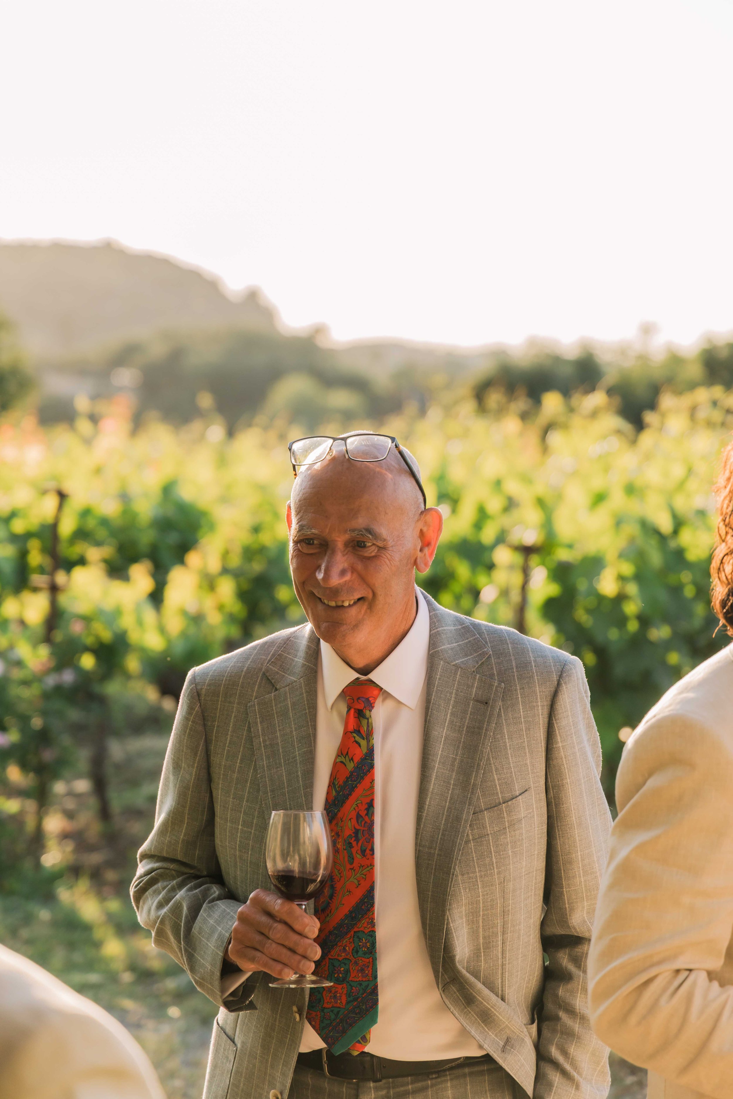 A man in a checked suit jacket and colorful tie holding a glass of red wine at an outdoor event, smiling, with a vineyard in the background.