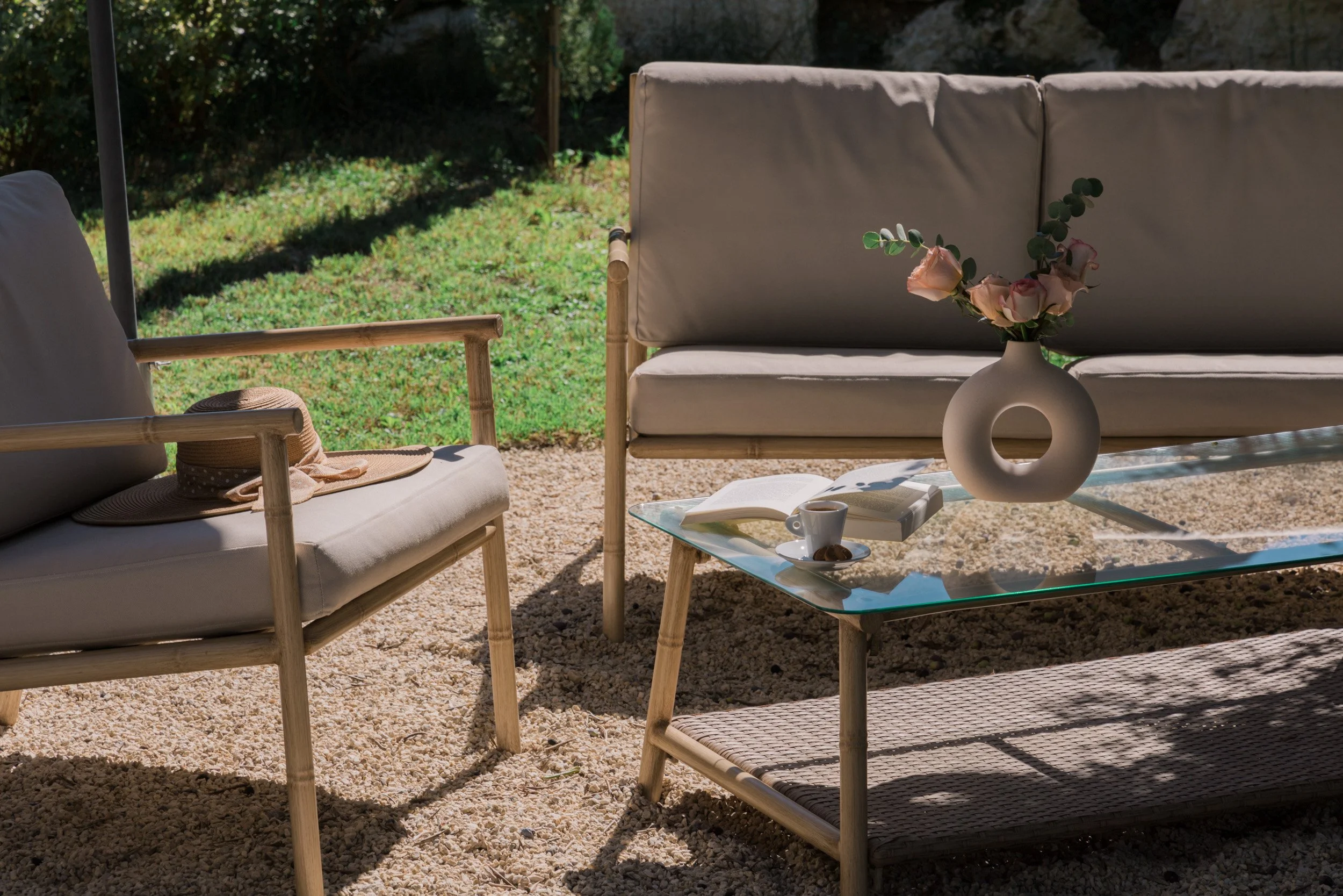 Outdoor patio furniture with a glass-top coffee table, a beige sofa, and a wooden armchair with cushions, all on a gravel surface under sunlight, with a backyard in the background. The coffee table has a white vase with pink flowers, an open book, a 