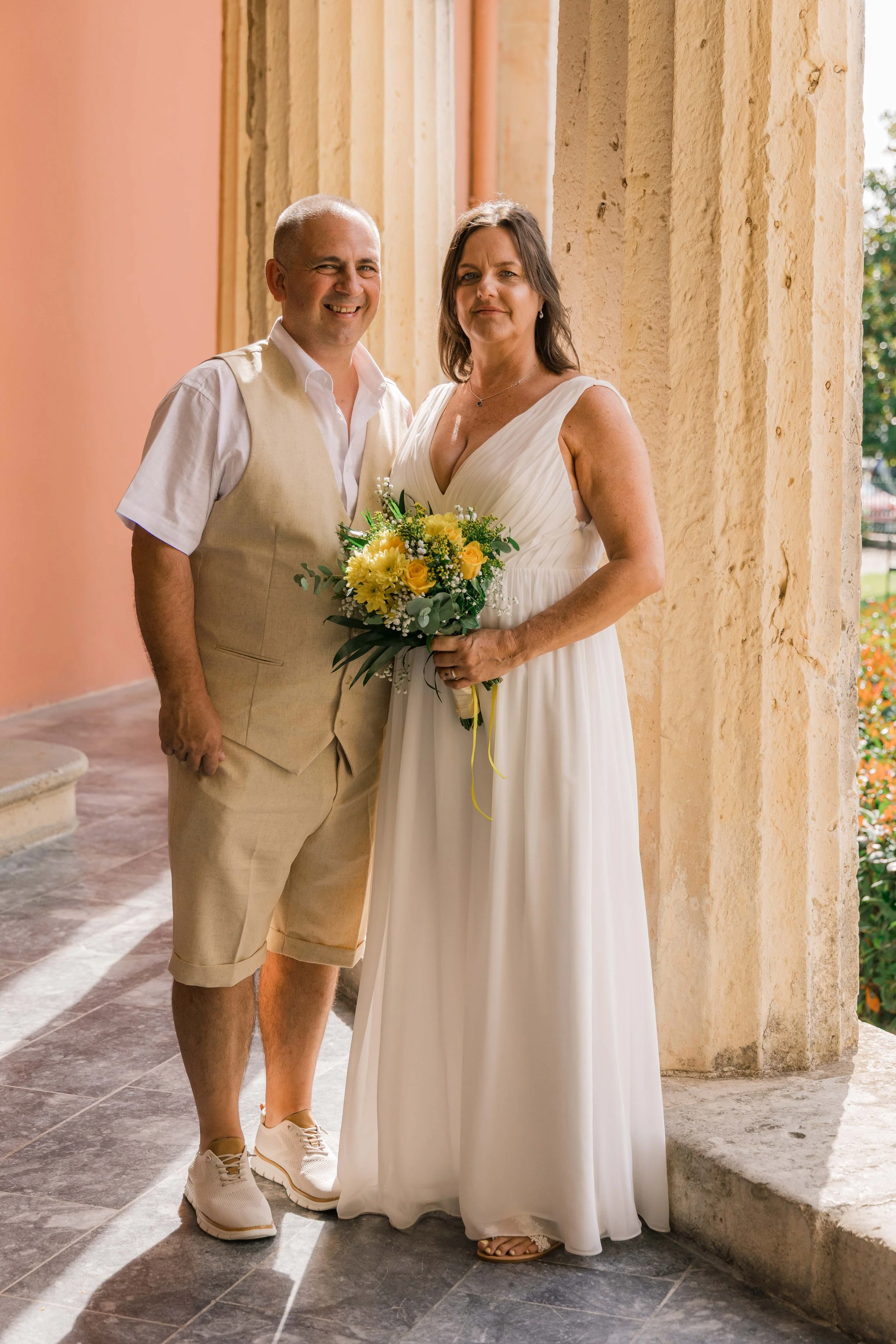 A couple standing together outside a building with stone columns and a pink wall. The woman is wearing a white dress and holding a bouquet of yellow flowers. The man is dressed casually with a beige vest and shorts.