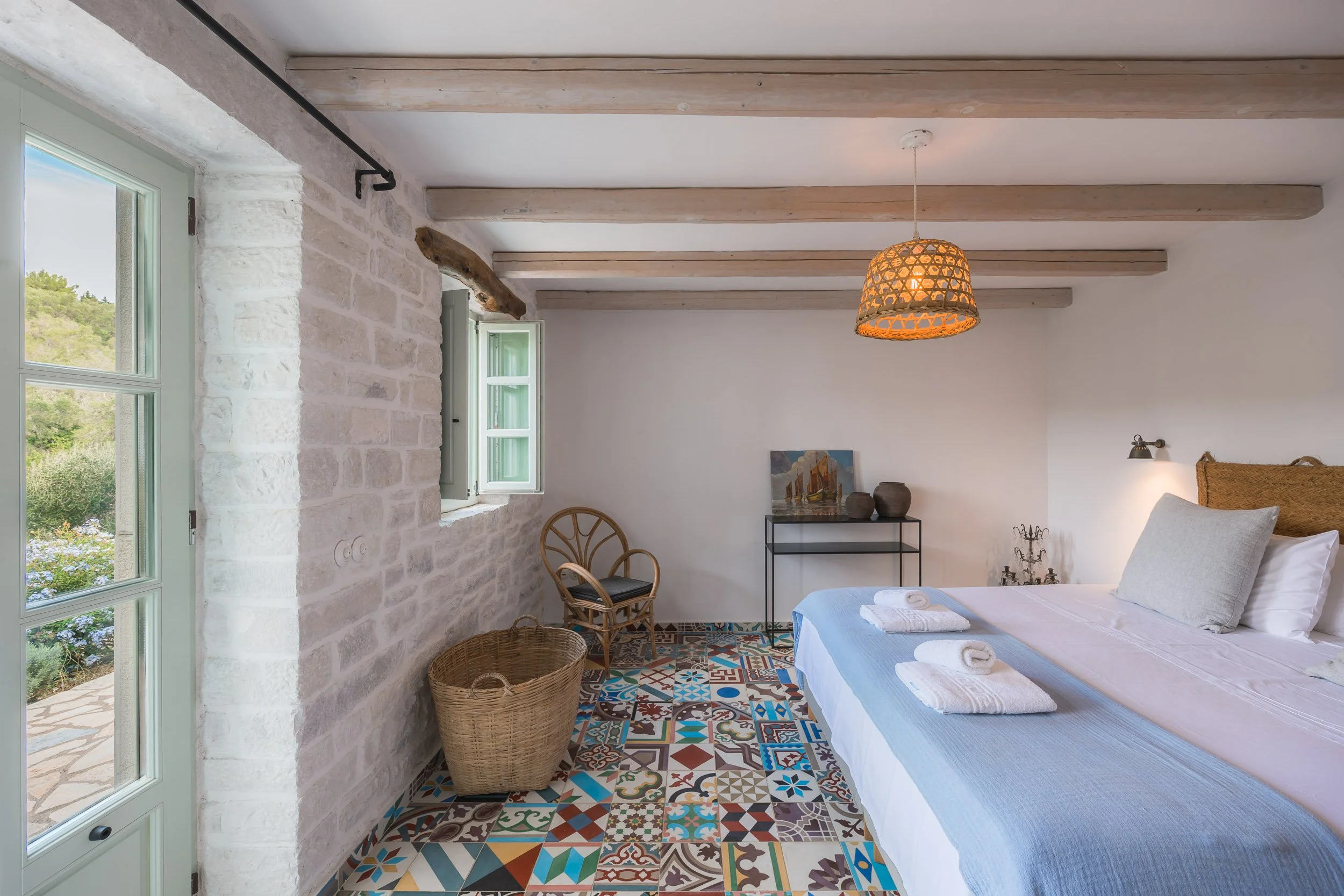 Cozy bedroom with white walls, exposed wooden ceiling beams, colorful patterned tile floor, bed with white linens and pillows, towels, wicker basket, wooden chair, small black metal table with artwork and pottery, open window with green shutters, han