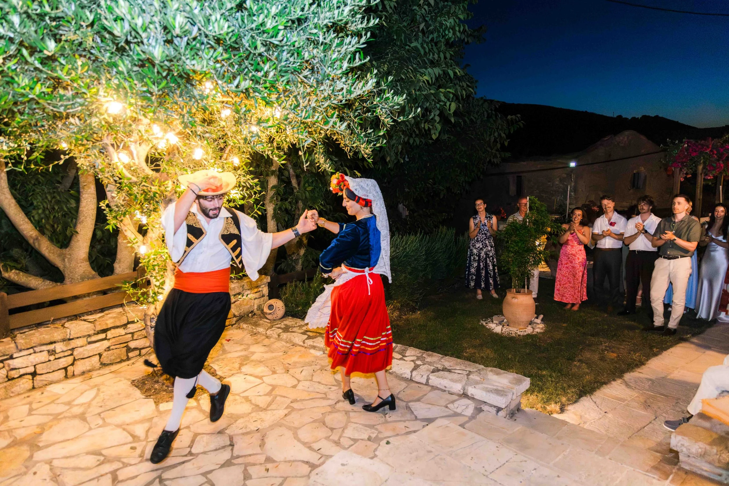 People dancing in traditional Greek attire at an outdoor celebration during the evening, with a group of onlookers clapping and smiling in the background.