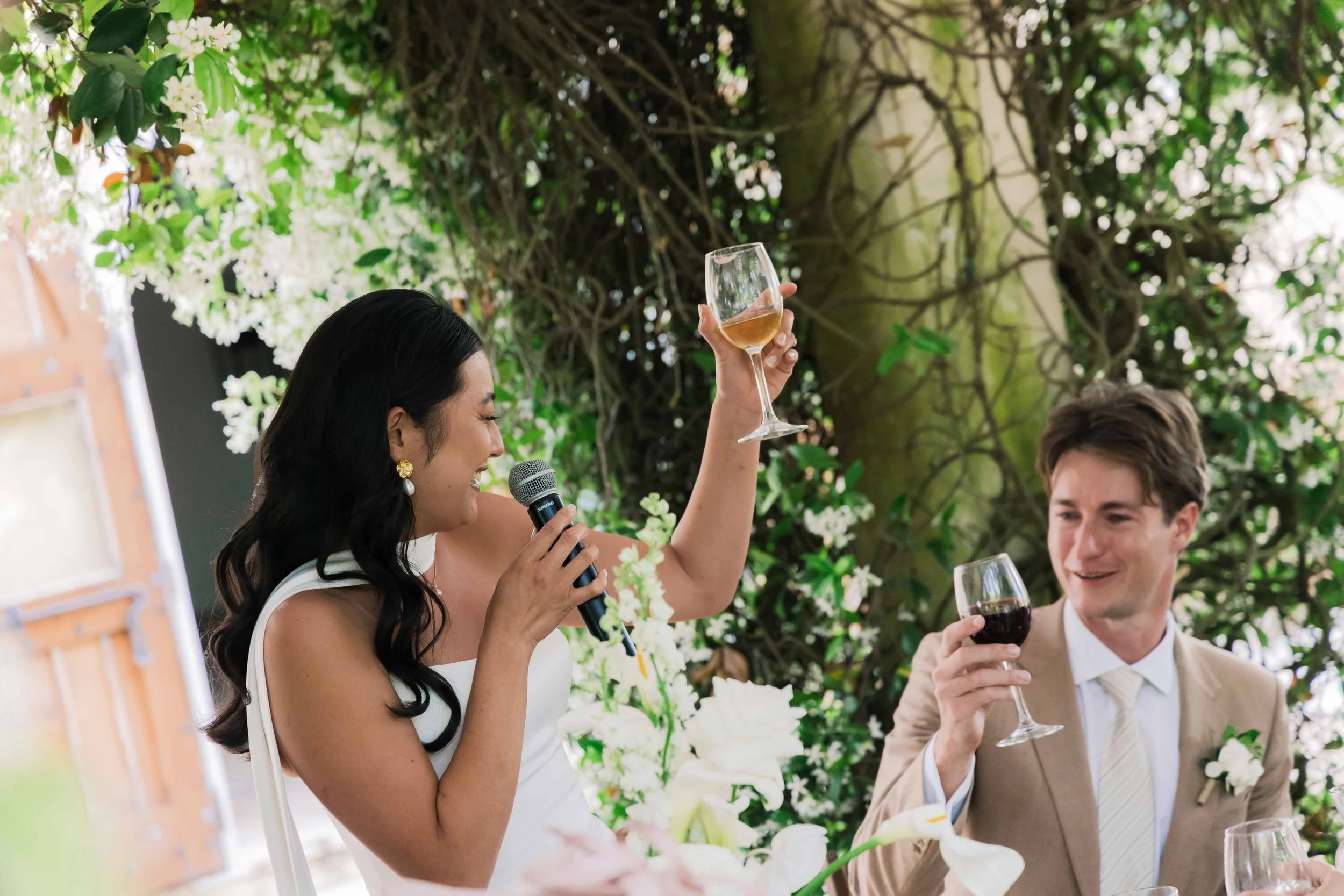 A woman in a white dress giving a speech with a microphone, holding a glass of rosé wine, at a wedding reception, with a man in a tan suit sitting nearby drinking red wine, both surrounded by white flowers and greenery.