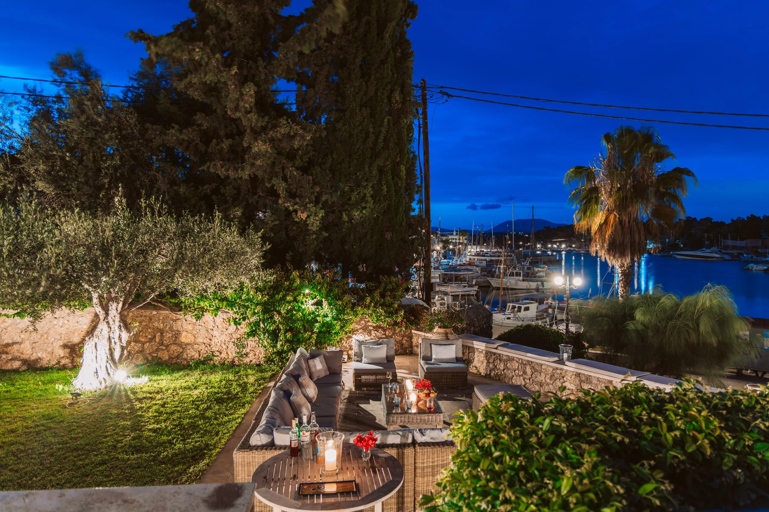 Outdoor patio at night overlooking a marina with boats, illuminated seating area with candles, flowers, trees, and bushes.