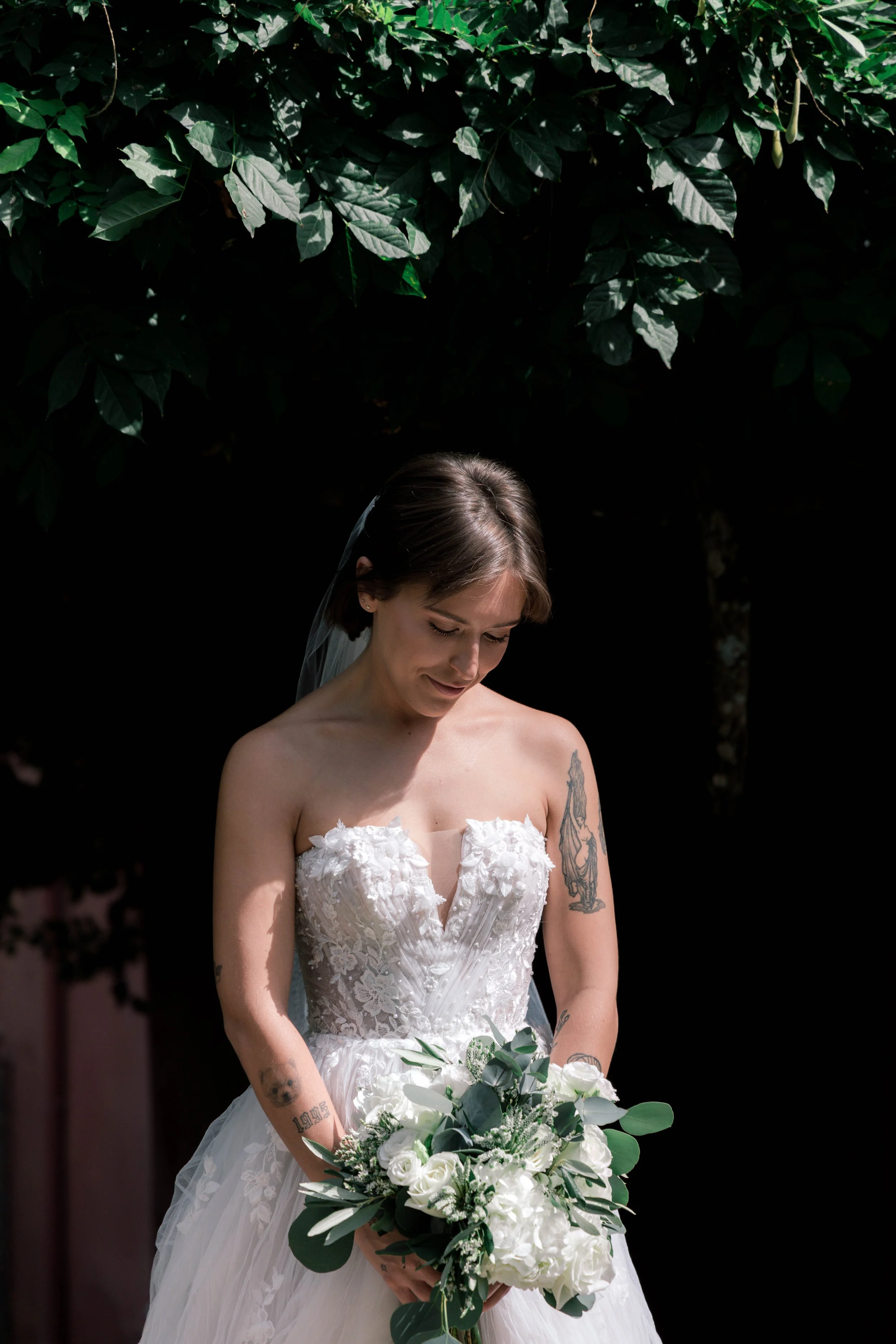 Bride in a strapless white wedding dress holding a bouquet of white roses and greenery, with tattoos on her arms, standing under a tree with green leaves.