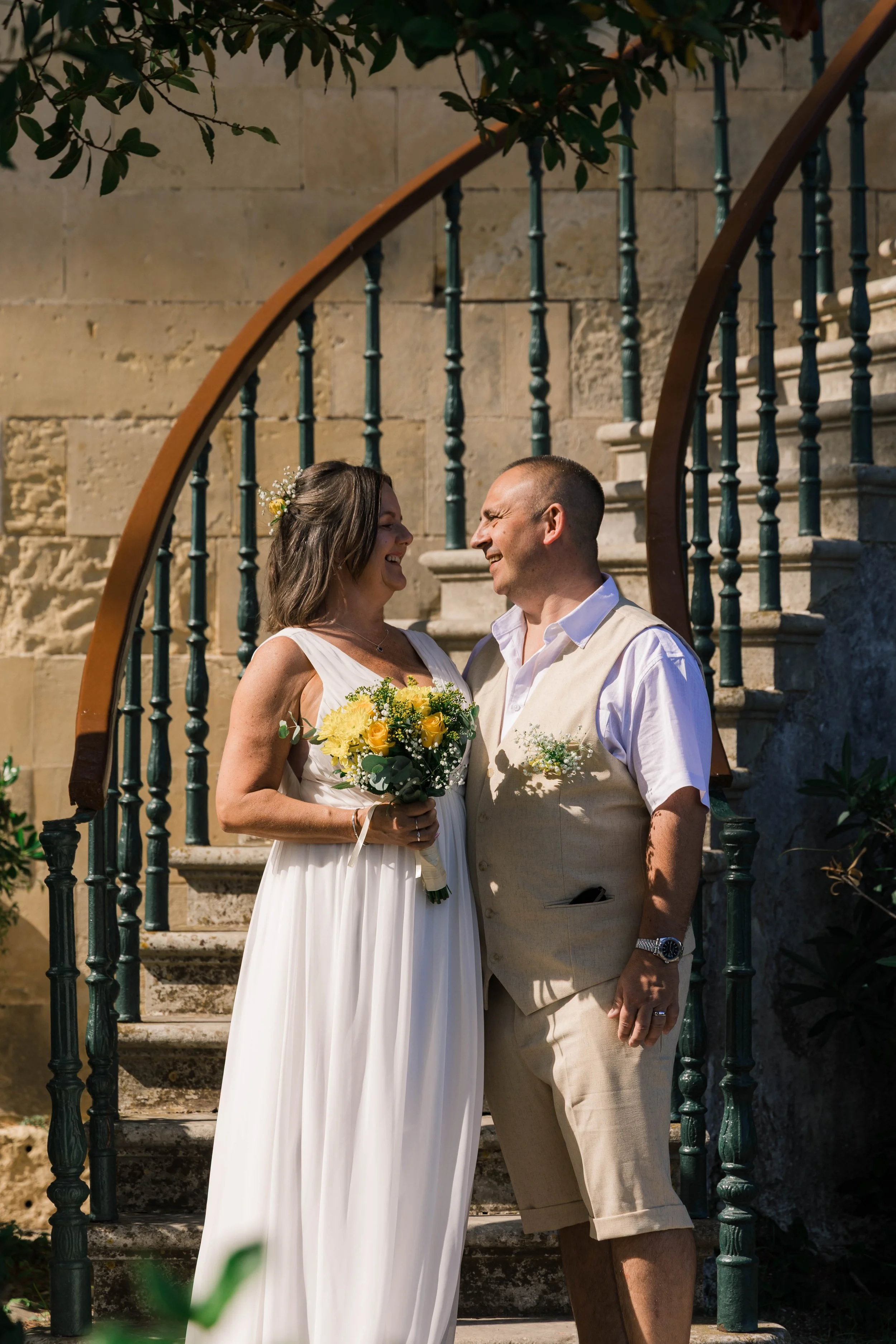 A bride and groom smiling at each other on an outdoor stone staircase, with the bride holding a yellow bouquet and wearing a white dress, and the groom in a light-colored vest and shorts.
