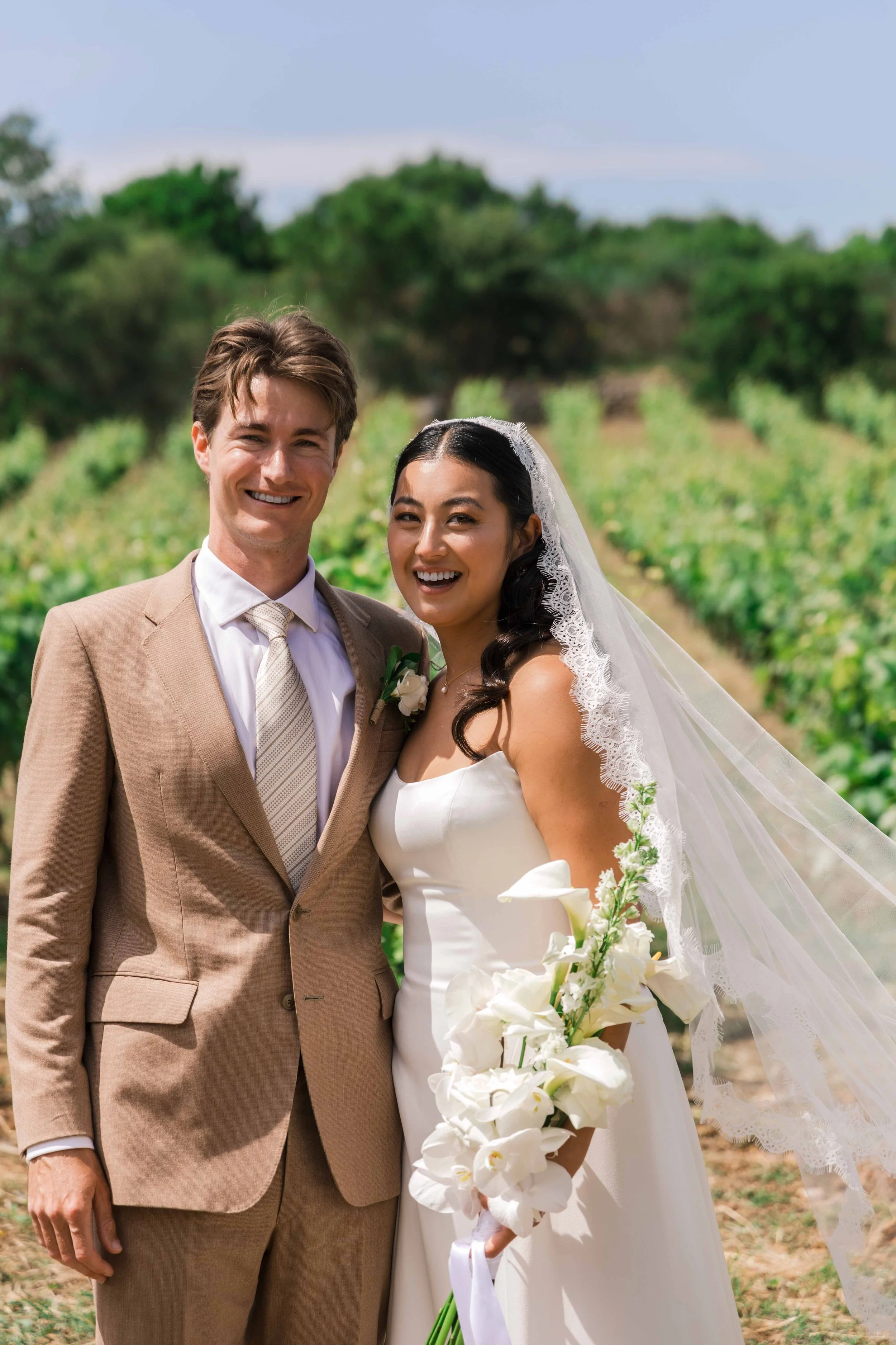 A happy bride and groom standing together outdoors on their wedding day, smiling, with a green field in the background. The bride is holding a bouquet of white flowers, and the groom is wearing a tan suit with a white shirt and tie.