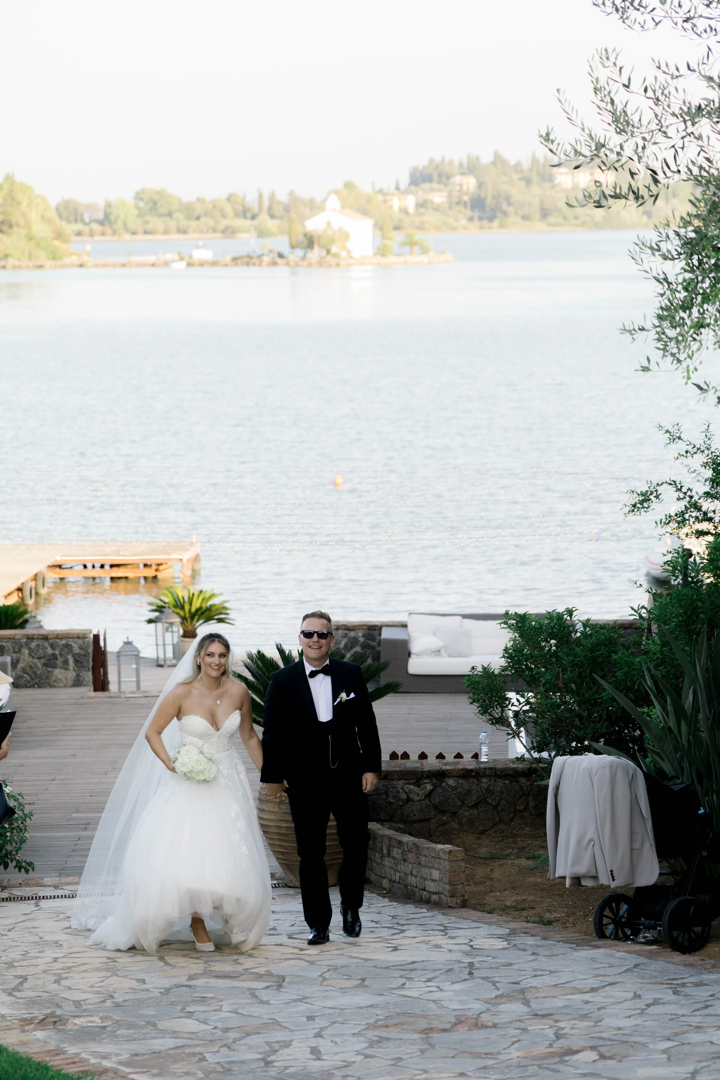 A newlywed couple walking outdoors beside a lake, with the bride in a white wedding gown holding a bouquet and the groom in a black tuxedo with sunglasses.