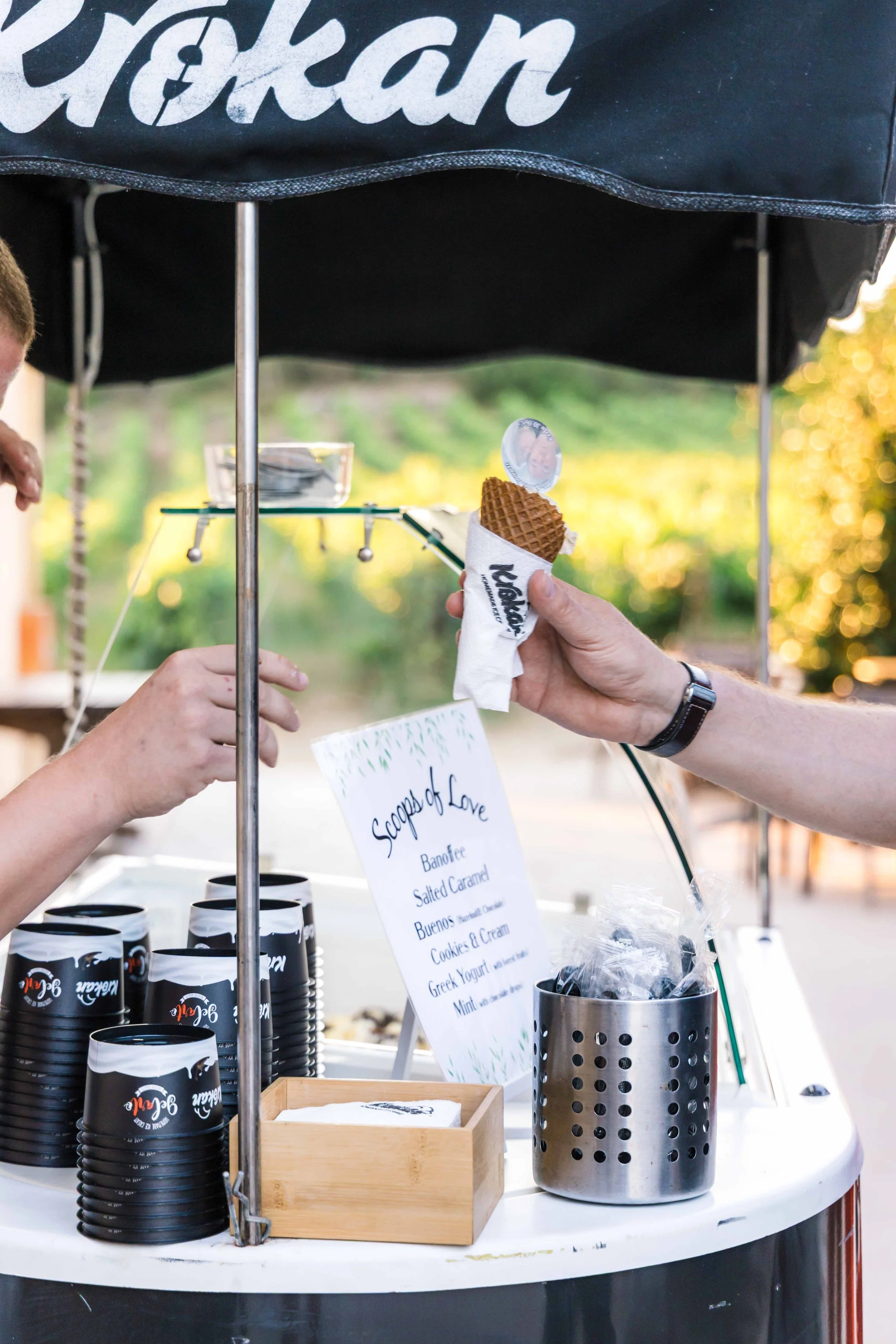 A person holding an ice cream cone at a street vendor stand, with stacks of black coffee cups and a sign listing flavors, under a black umbrella with white text.