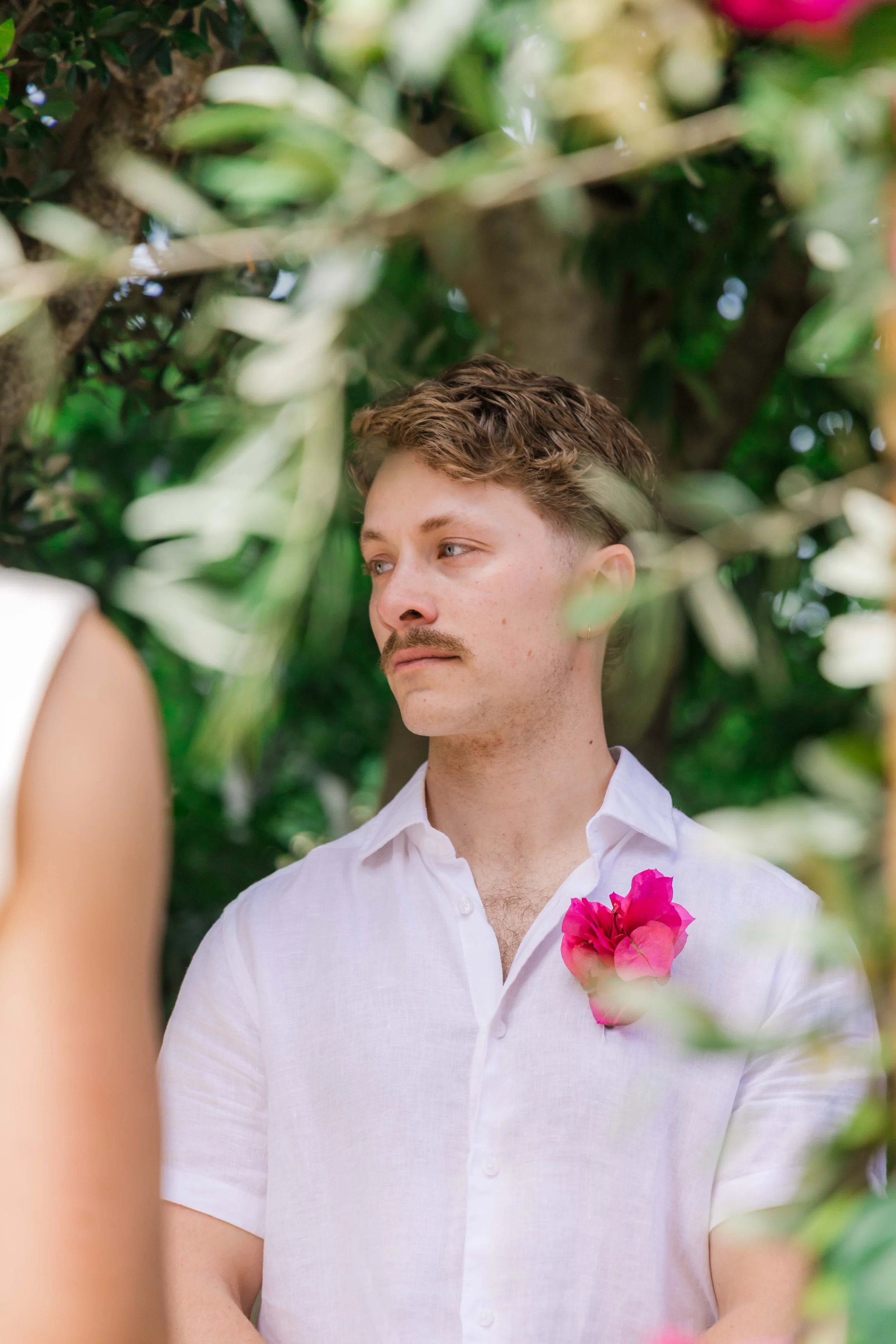 A man with light skin, a mustache, and light brown hair stands outdoors among green foliage, wearing a white shirt with a pink flower boutonniere on his chest, looking to the side.