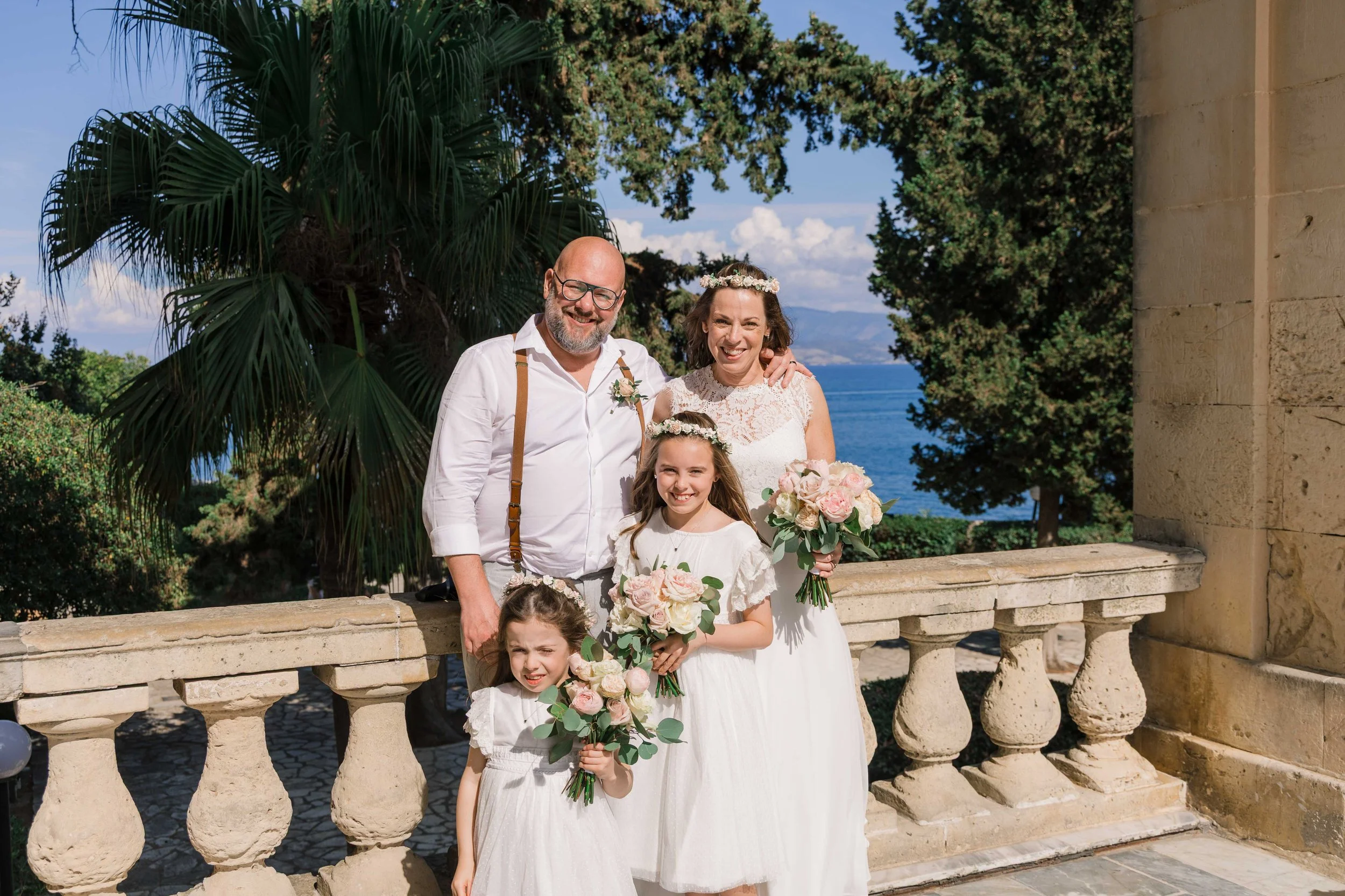 A family celebrating a wedding outdoors near the ocean, with a man, woman, and two young girls dressed in white, holding bouquets of pink and white flowers, standing on a stone balcony with palm trees and a scenic view in the background.
