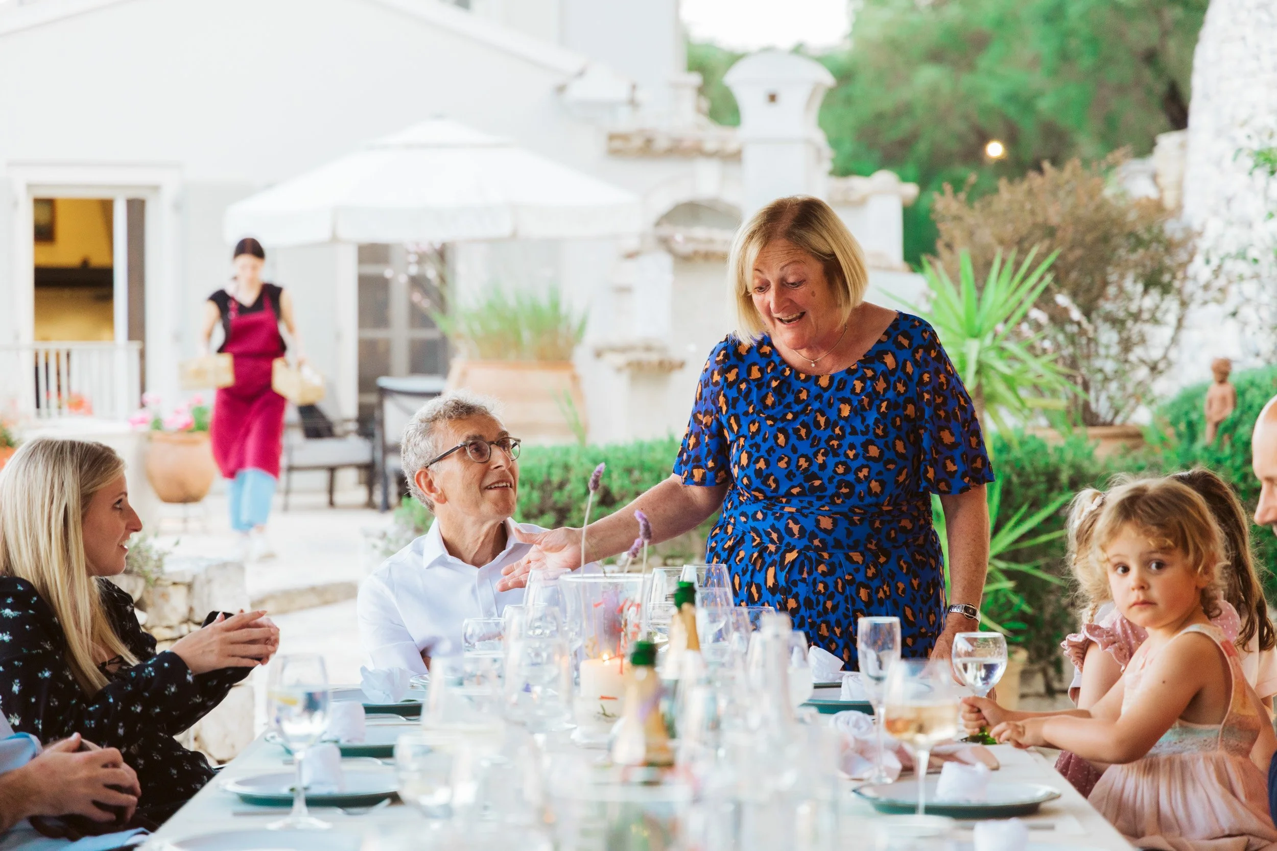 A family gathering at an outdoor celebration with a woman in a blue leopard print dress talking to a seated elderly man, surrounded by children and adults at a table with drinks and decorations, in a garden setting with greenery and a white building 