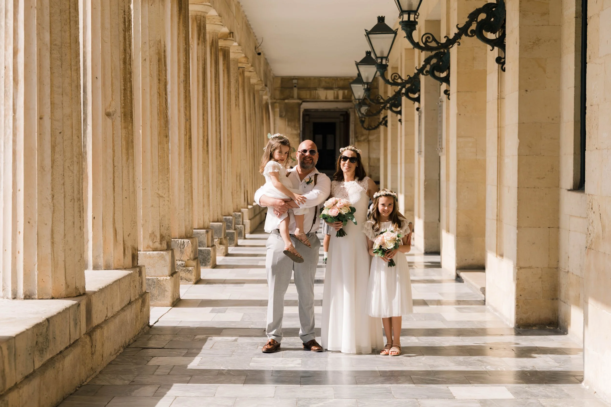 A family dressed in wedding attire standing in a sunny corridor with stone columns and vintage lamps, holding bouquets of flowers.