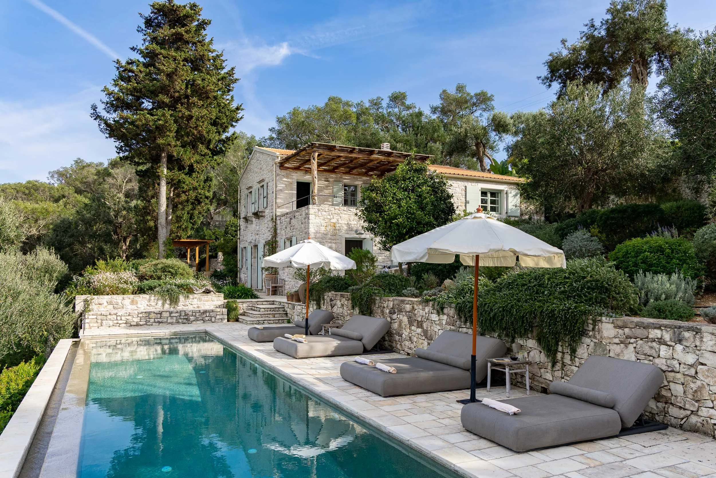 Luxury outdoor pool area with lounge chairs, umbrellas, and a stone house surrounded by greenery and trees.