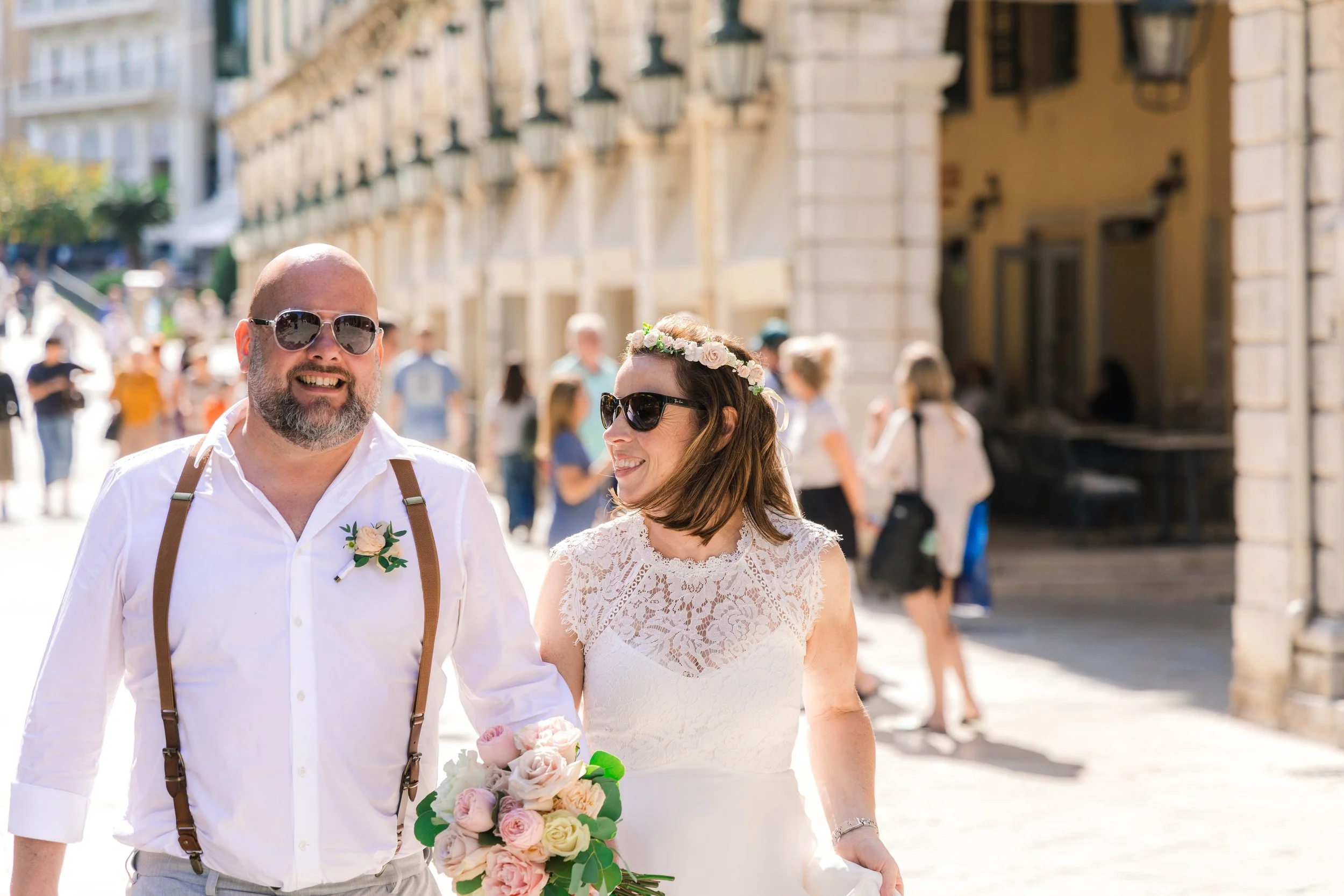 A smiling woman in a white lace wedding dress with a floral crown holding a bouquet, walking arm-in-arm with a bearded man in a white shirt, suspenders, and sunglasses on a sunny street with people and historic buildings in the background.