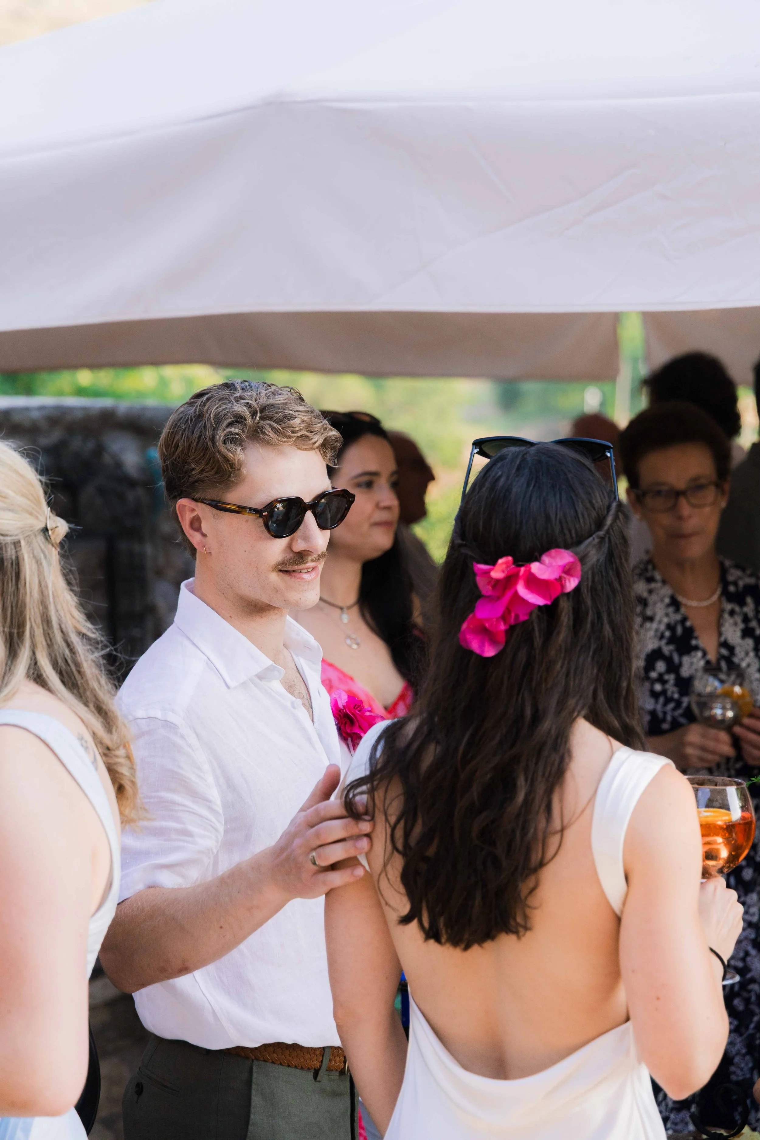 People gathered at an outdoor social event under a tent, including a man with glasses and a white shirt, women with floral accessories, and others holding drinks.