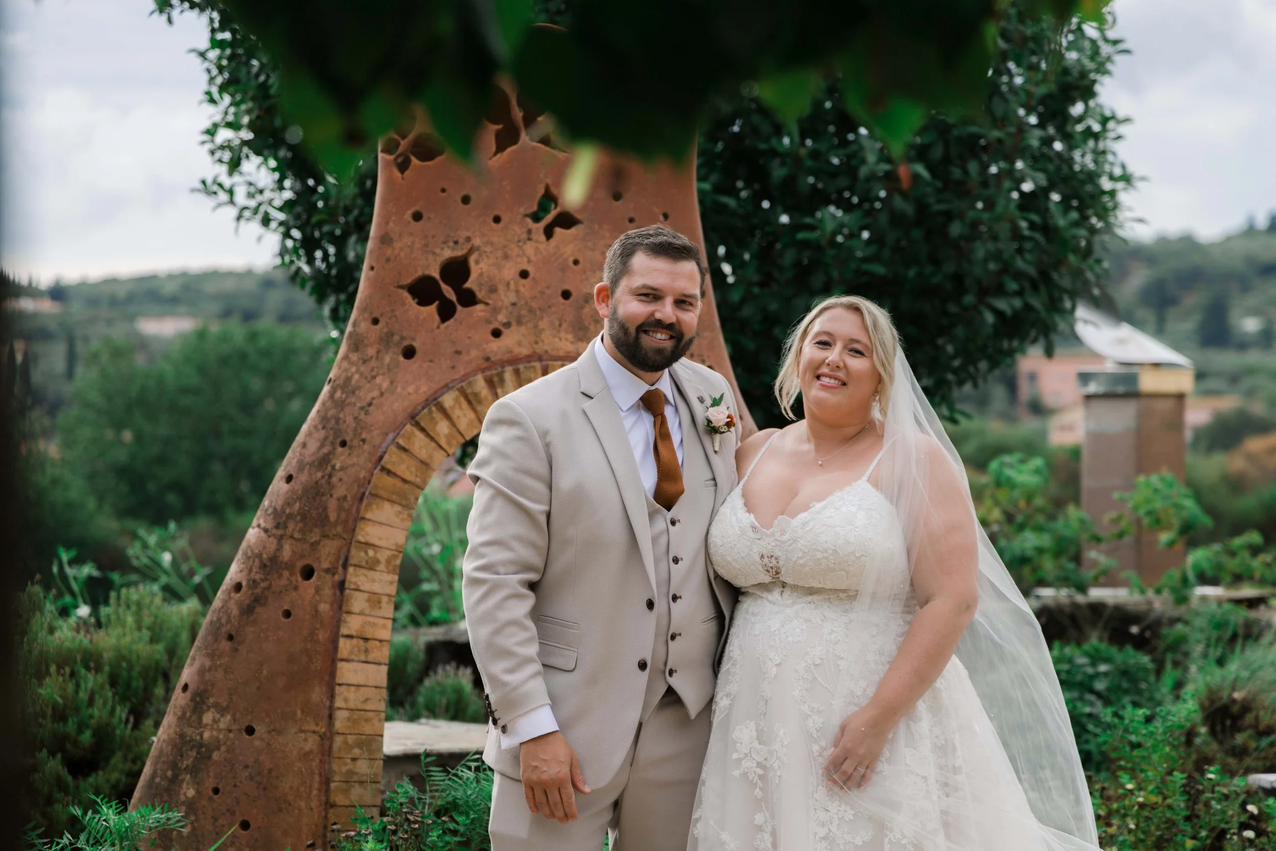 A bride and groom standing outdoors in front of a metal sculpture with butterflies, greenery, and cloudy sky in the background.