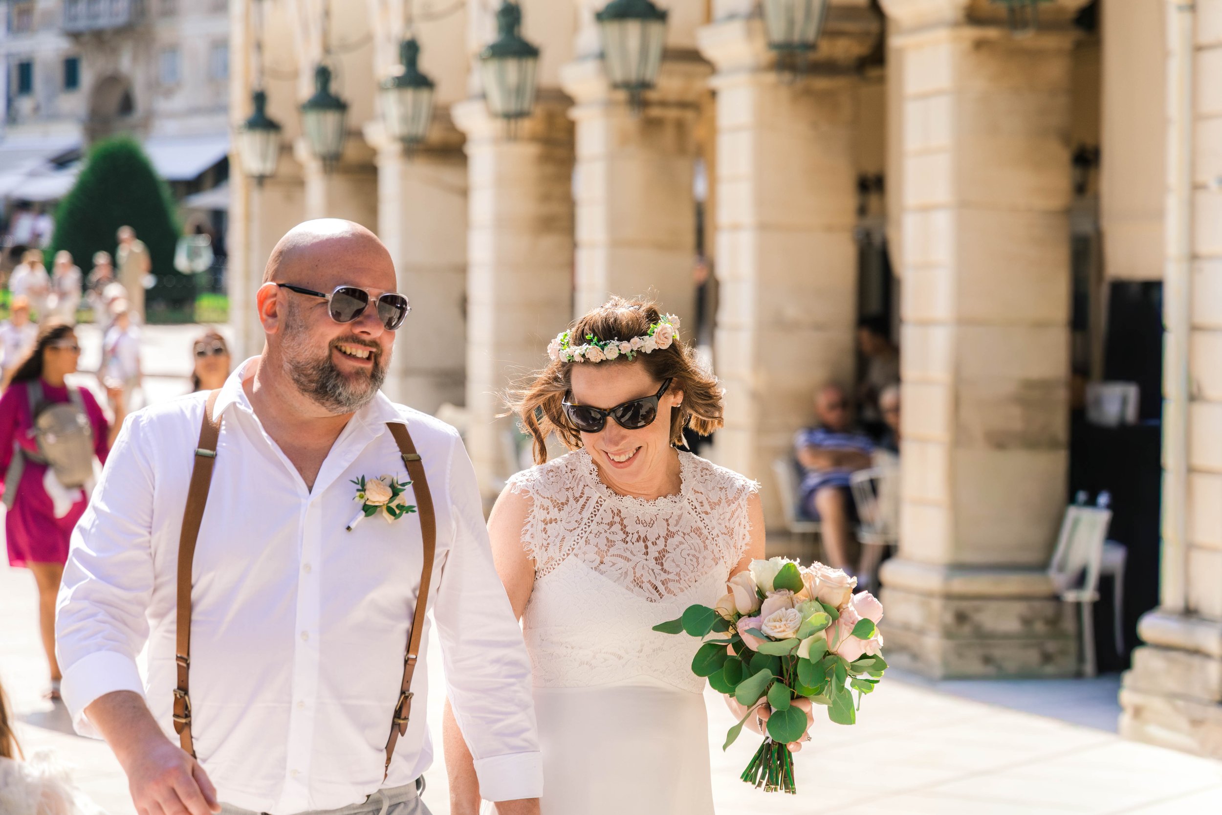 A smiling couple walking outdoors on a sunny day, with the woman in a wedding dress holding a bouquet of flowers, and the man in a white shirt with suspenders and sunglasses, surrounded by people and historic buildings.