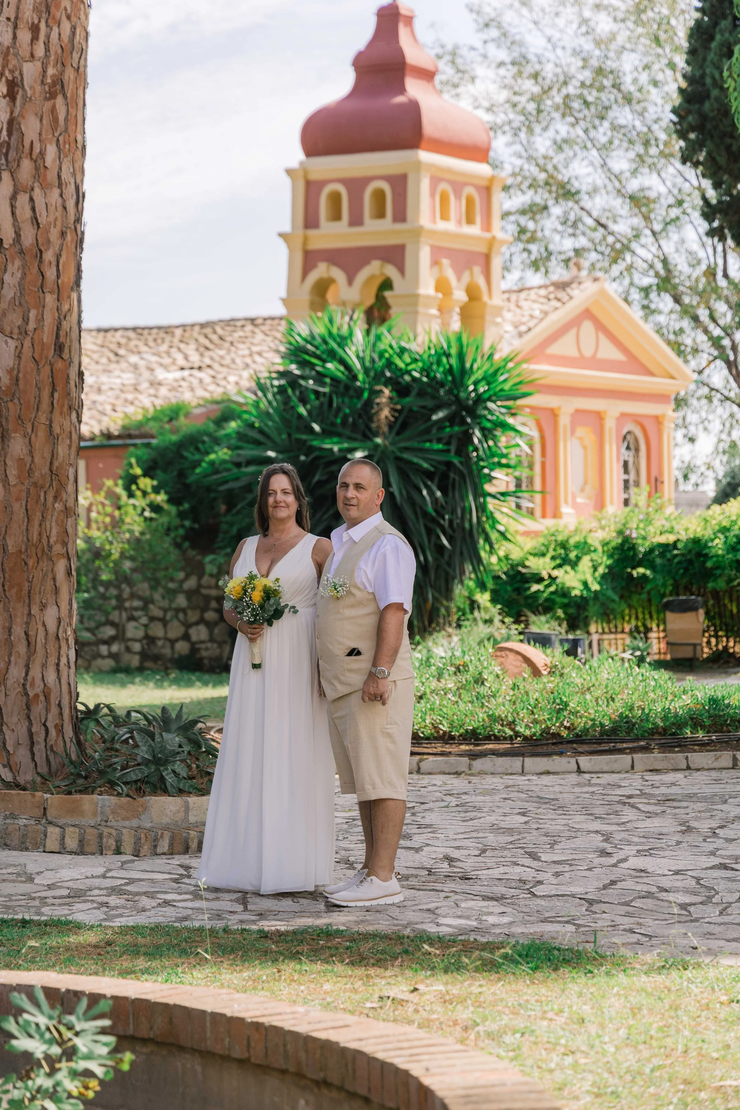 A couple dressed in wedding attire standing outdoors in front of a colorful church, with greenery and trees in the background.