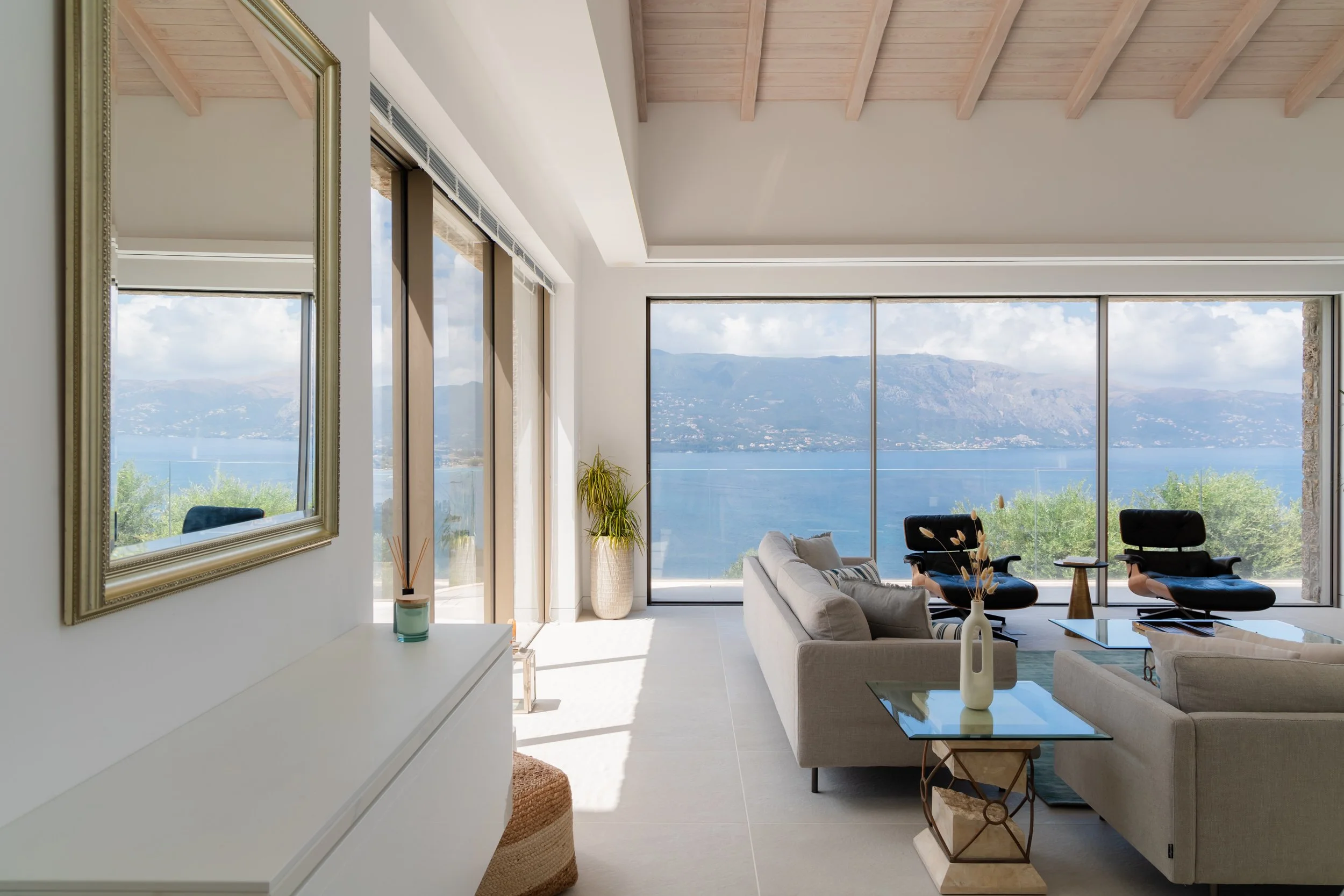 Living room with large windows overlooking a lake and mountains, modern furniture, beige sofa, black chairs, and decorative items.