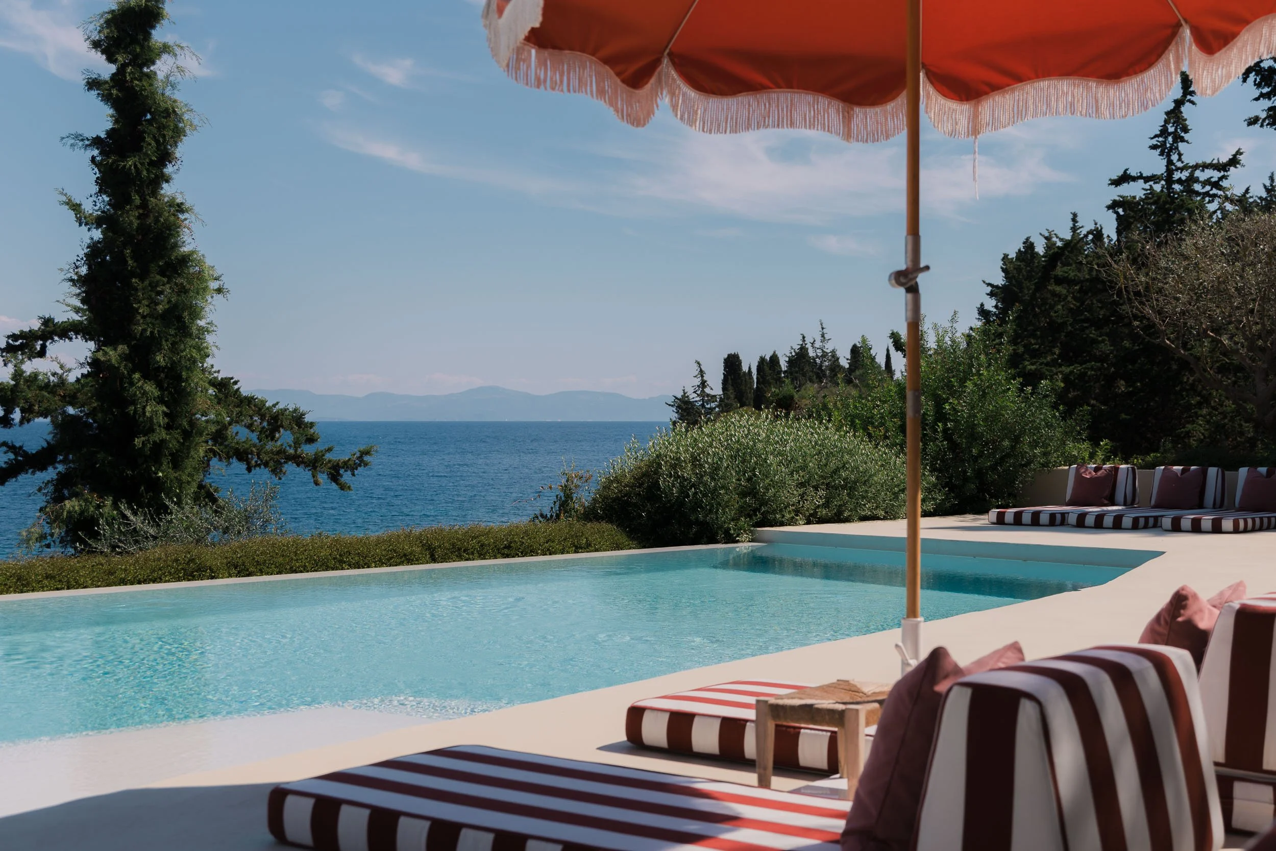 Poolside lounge chairs with striped cushions and pillows under a large orange umbrella overlooking a swimming pool, with trees, bushes, and a body of water in the background under a blue sky.
