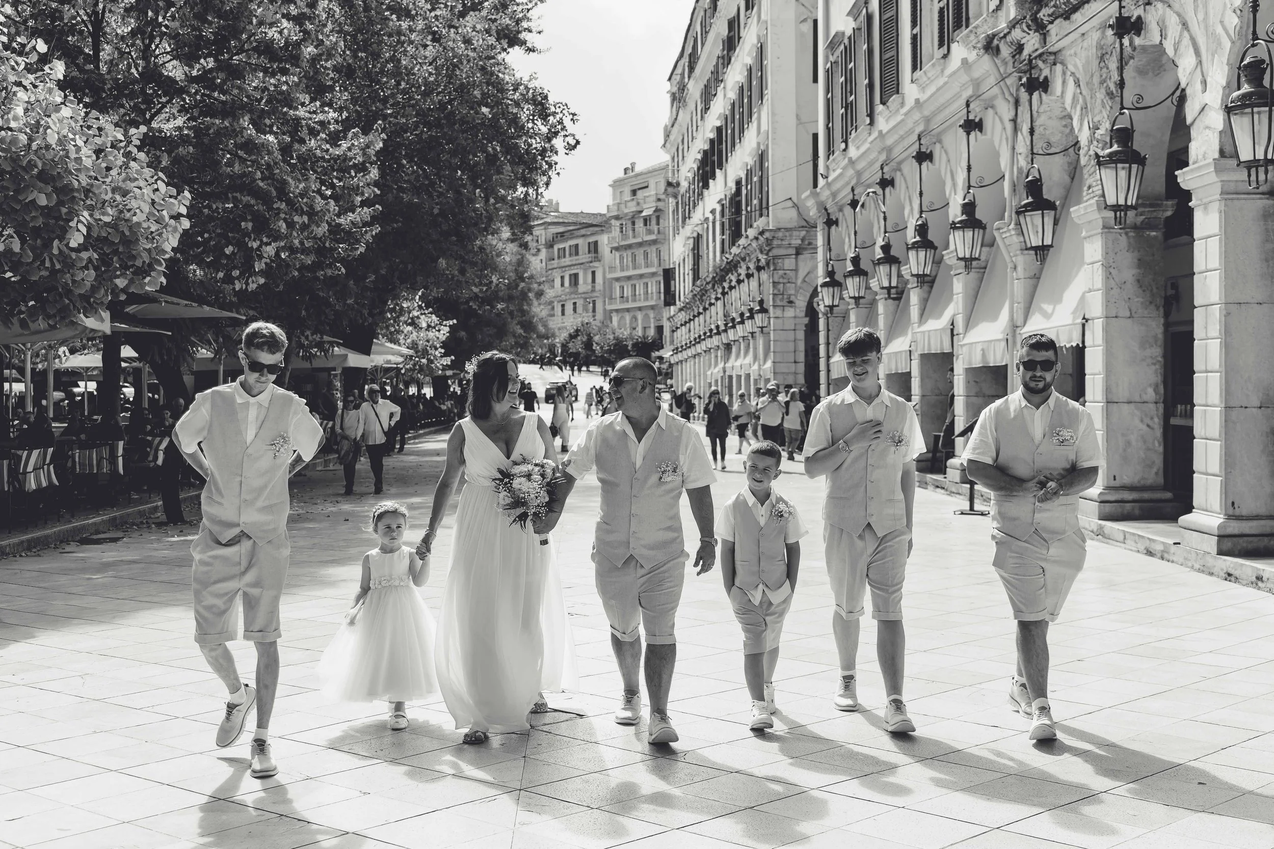Black and white photo of a wedding procession walking down a city street, including a bride, groom, children, and groomsmen, all dressed in light-colored attire.
