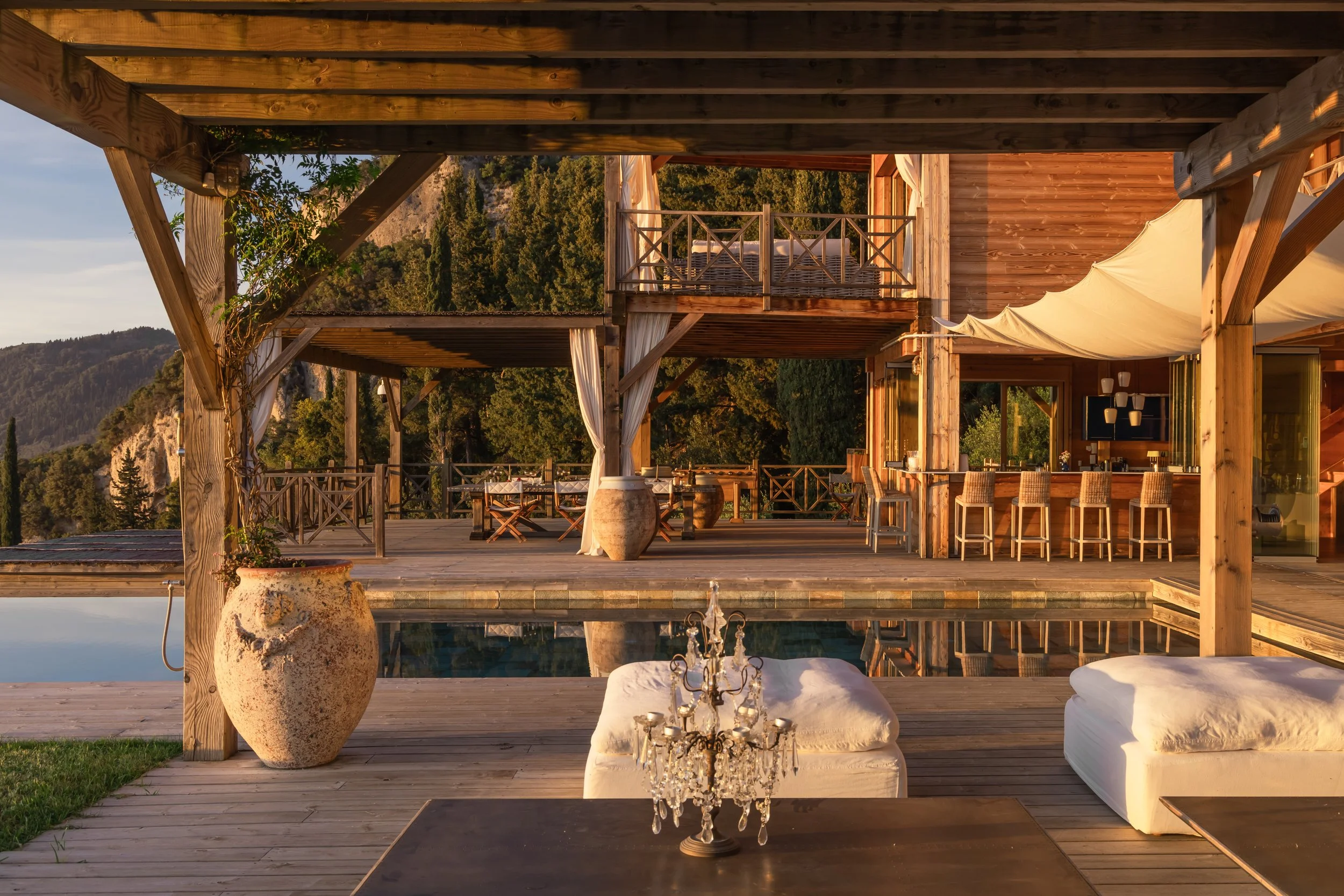 Outdoor wooden deck area with a pool, daytime, overlooking mountains, with chairs, tables, and a crystal chandelier on a table.