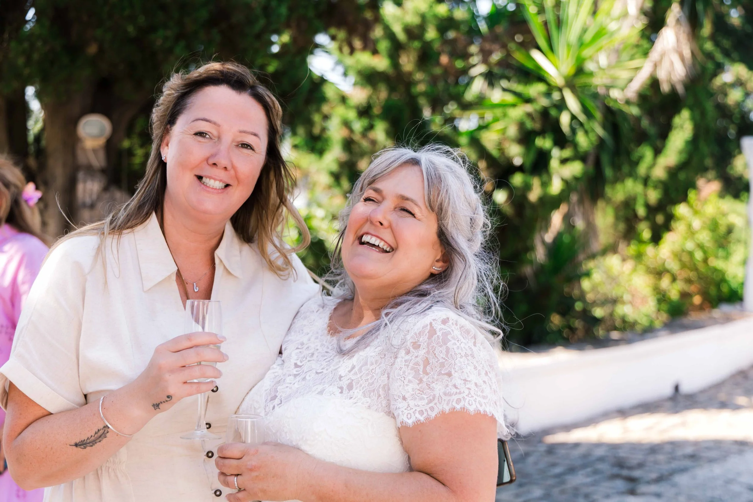 Two women smiling outdoors, holding glasses, one with a white lace top and the other with a light-colored button-up shirt, with greenery in the background.