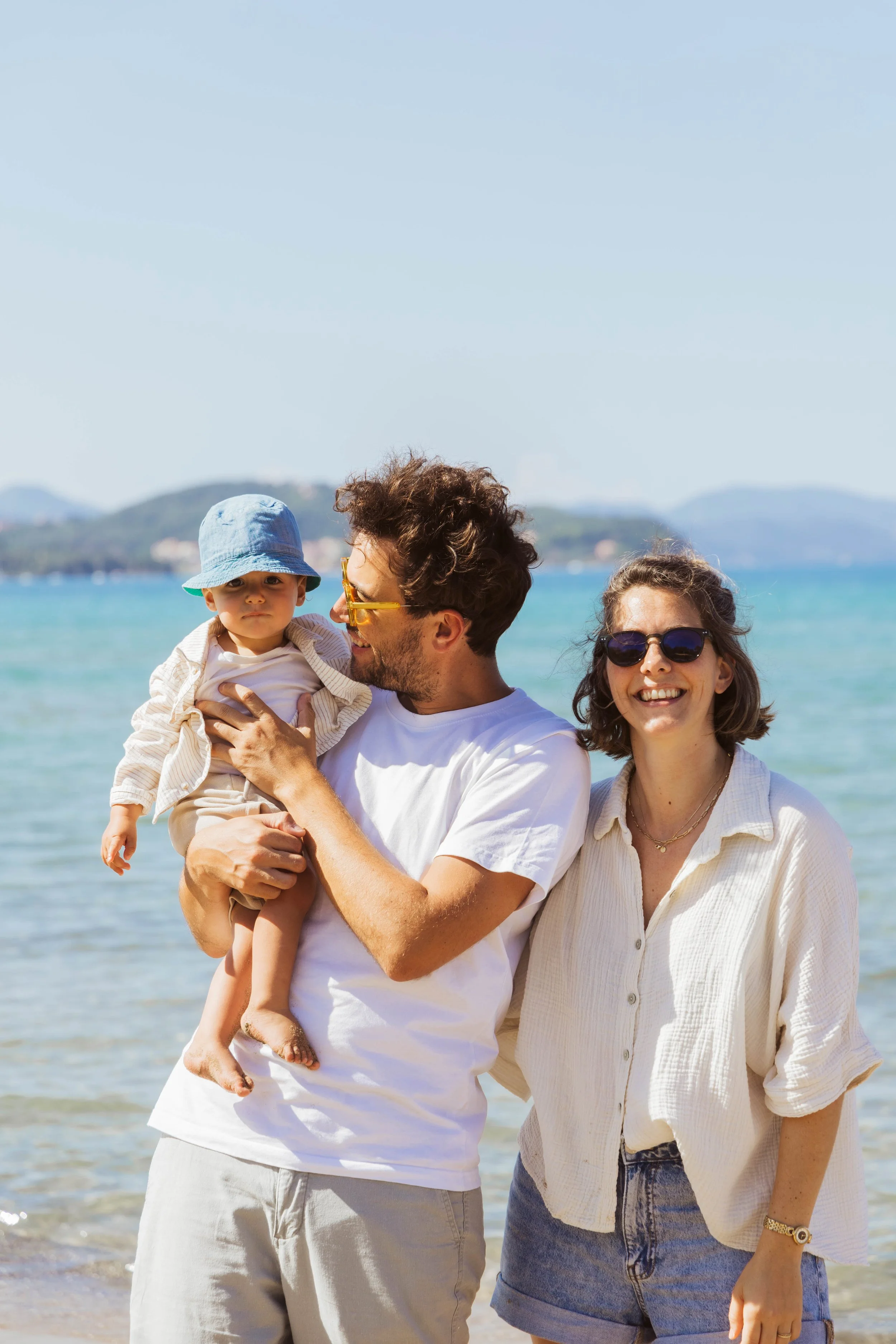 A smiling family of three at the beach, with a man holding a toddler boy, and a woman standing beside them, all in summer clothing.