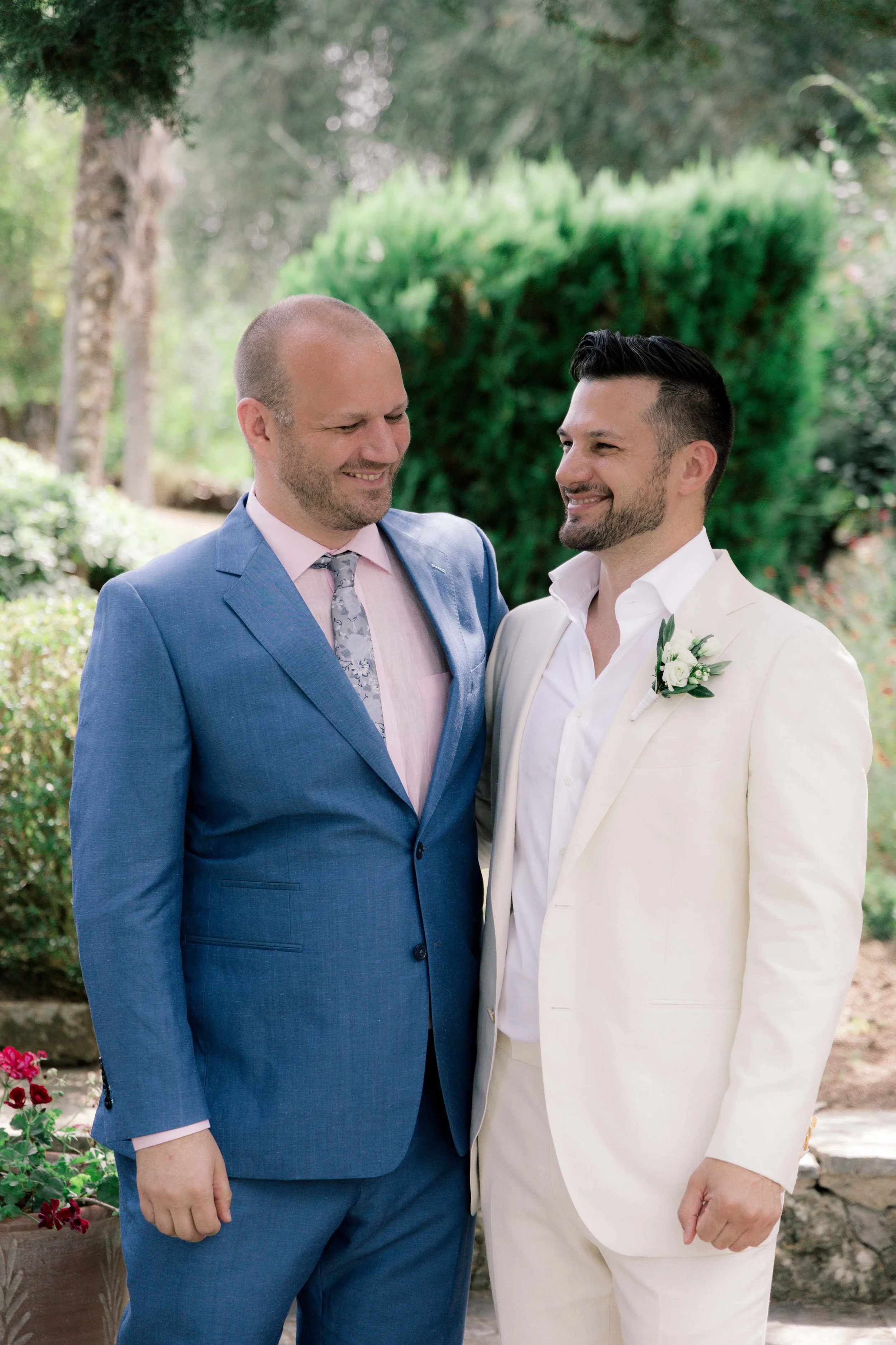 Two men in suits standing close together outdoors, smiling at each other during a wedding ceremony.