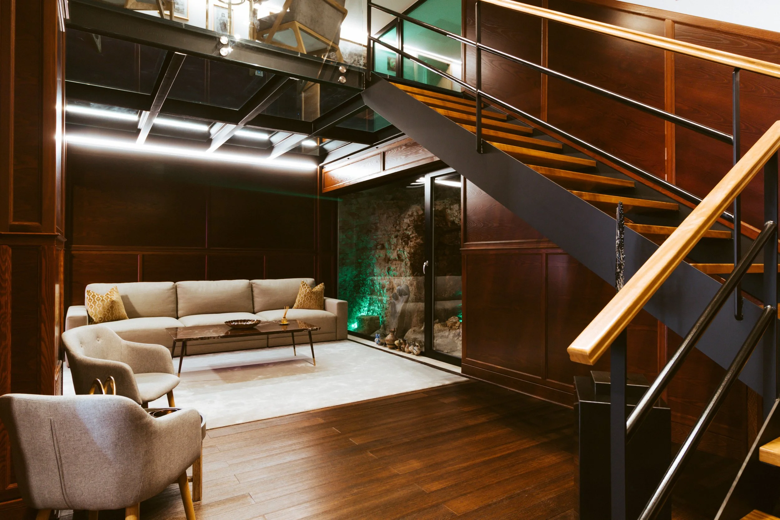 Interior view of a modern living room with wooden walls and flooring, beige sofa with yellow cushions, two armchairs, a glass coffee table, a white area rug, and a staircase with wooden steps and black railing.