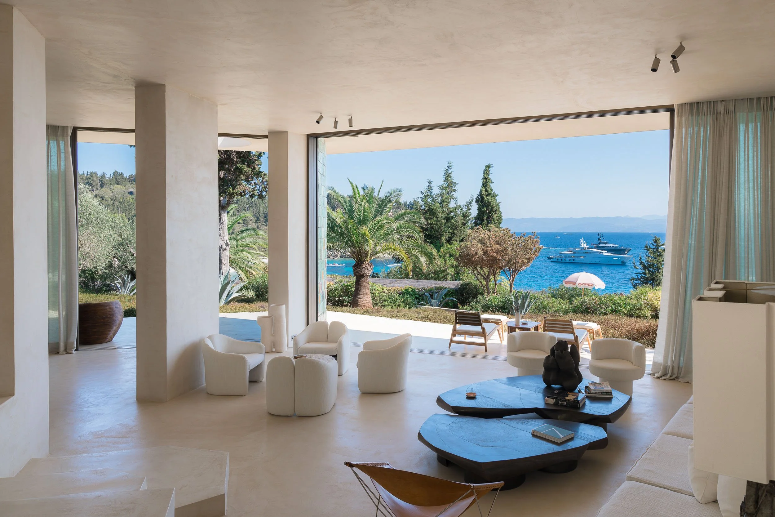 Living room with large open windows overlooking a seascape with boats, palm trees, and outdoor seating.