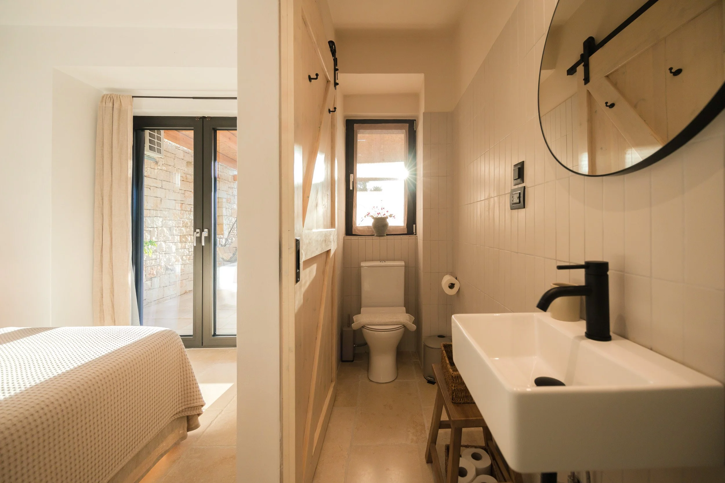 Small bathroom with a white toilet, a small white sink with a black faucet, a round mirror on the wall, and a window with sunlight shining through, next to a wooden sliding door.