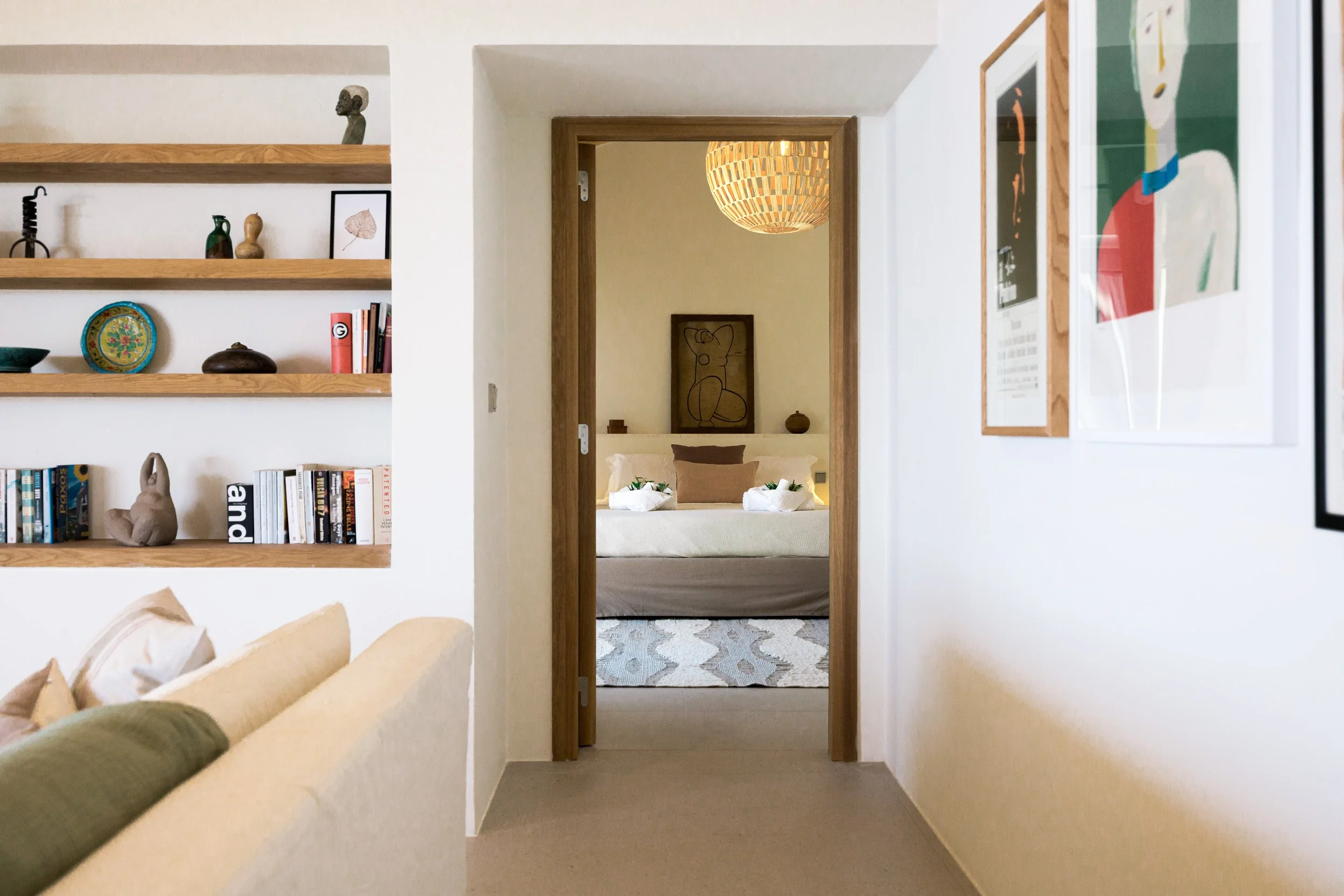 View through a doorway into a bedroom with a bed, pillows, and wall art, with a hallway on either side featuring framed pictures, a wooden shelf with decorative items, and part of a beige couch with cushions in the foreground.