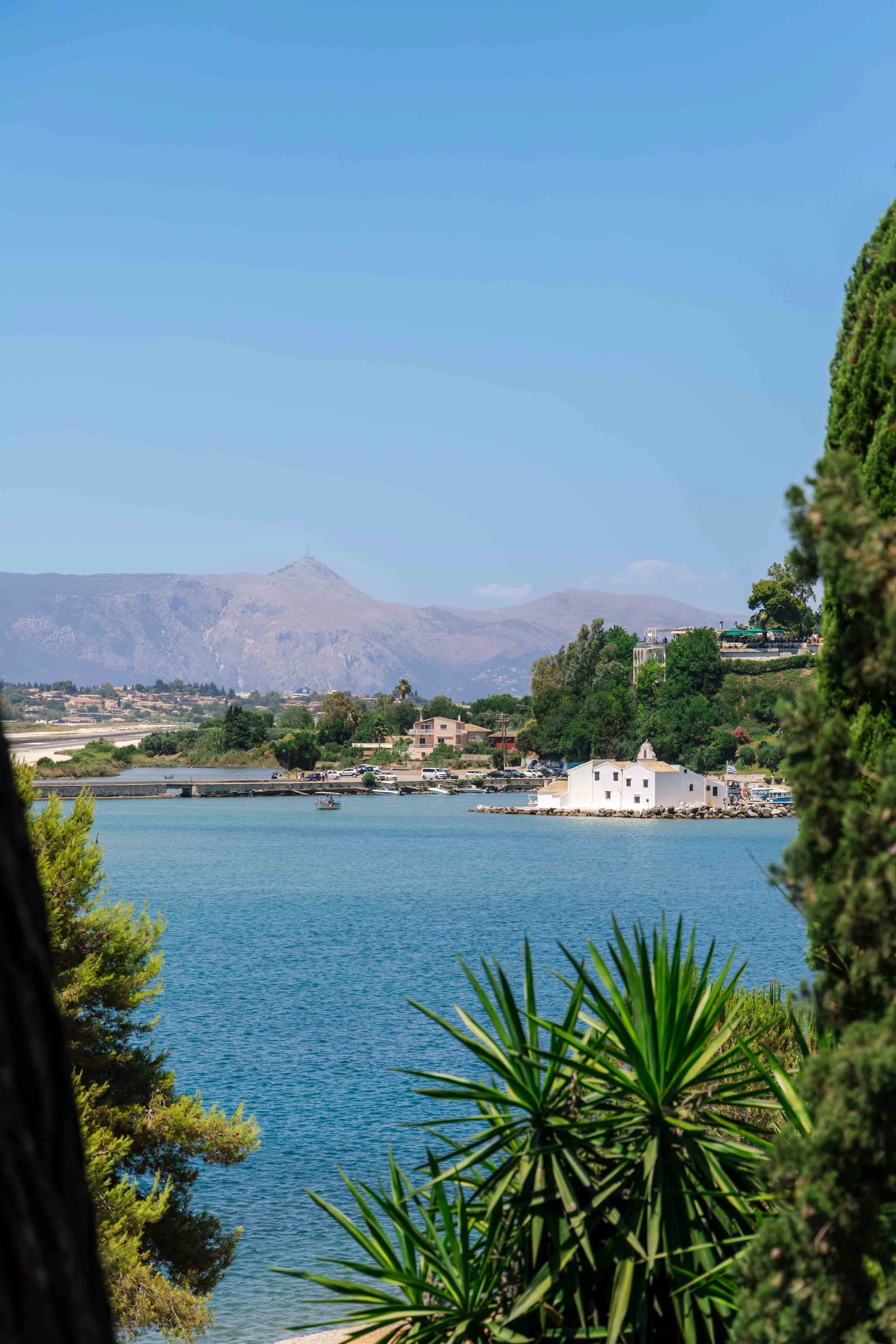 Scenic view of a water body with small buildings on the shoreline, surrounded by lush greenery and mountains in the background under a clear blue sky.
