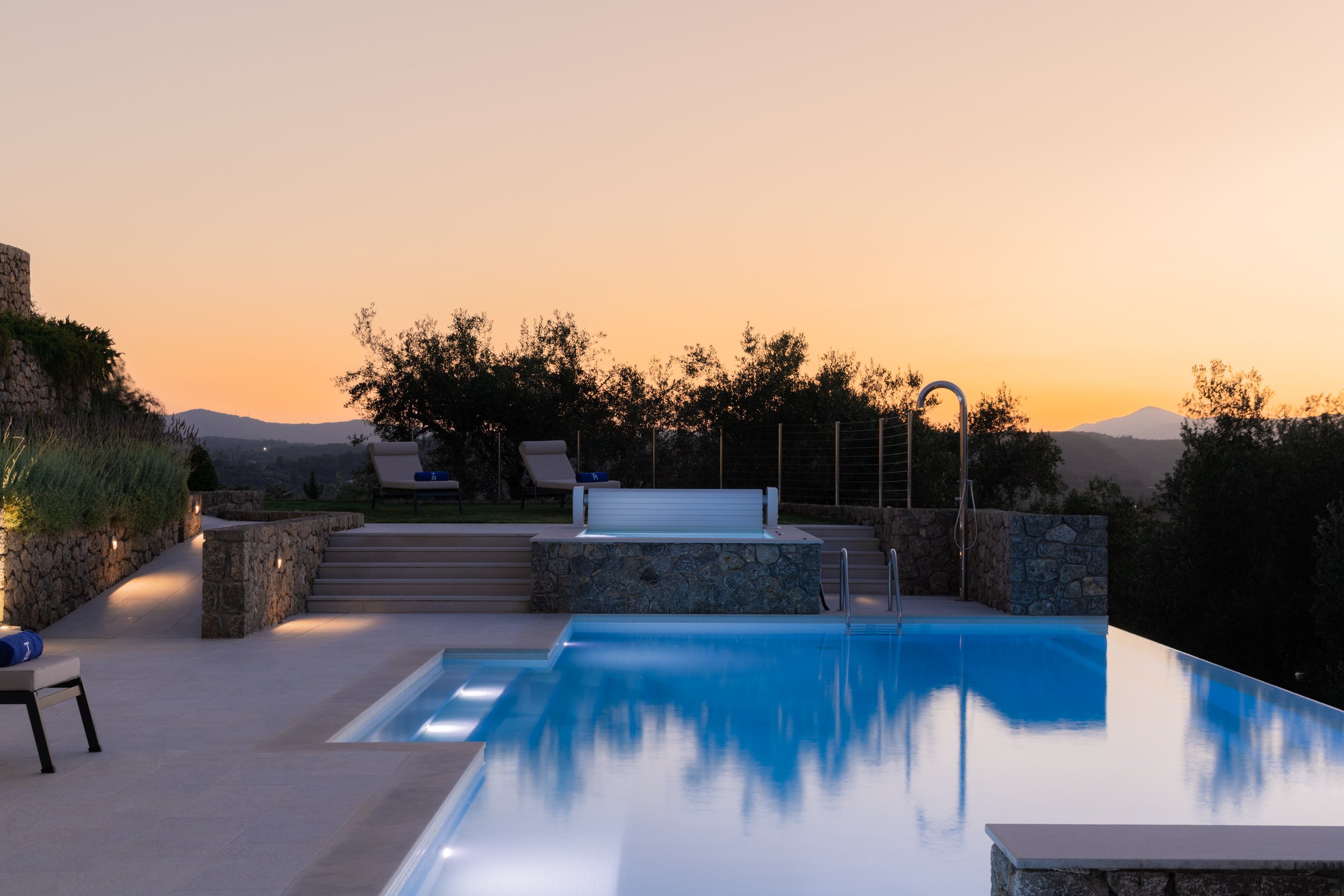 An outdoor swimming pool at sunset with lounge chairs and a hot tub on a terrace surrounded by trees and hills.