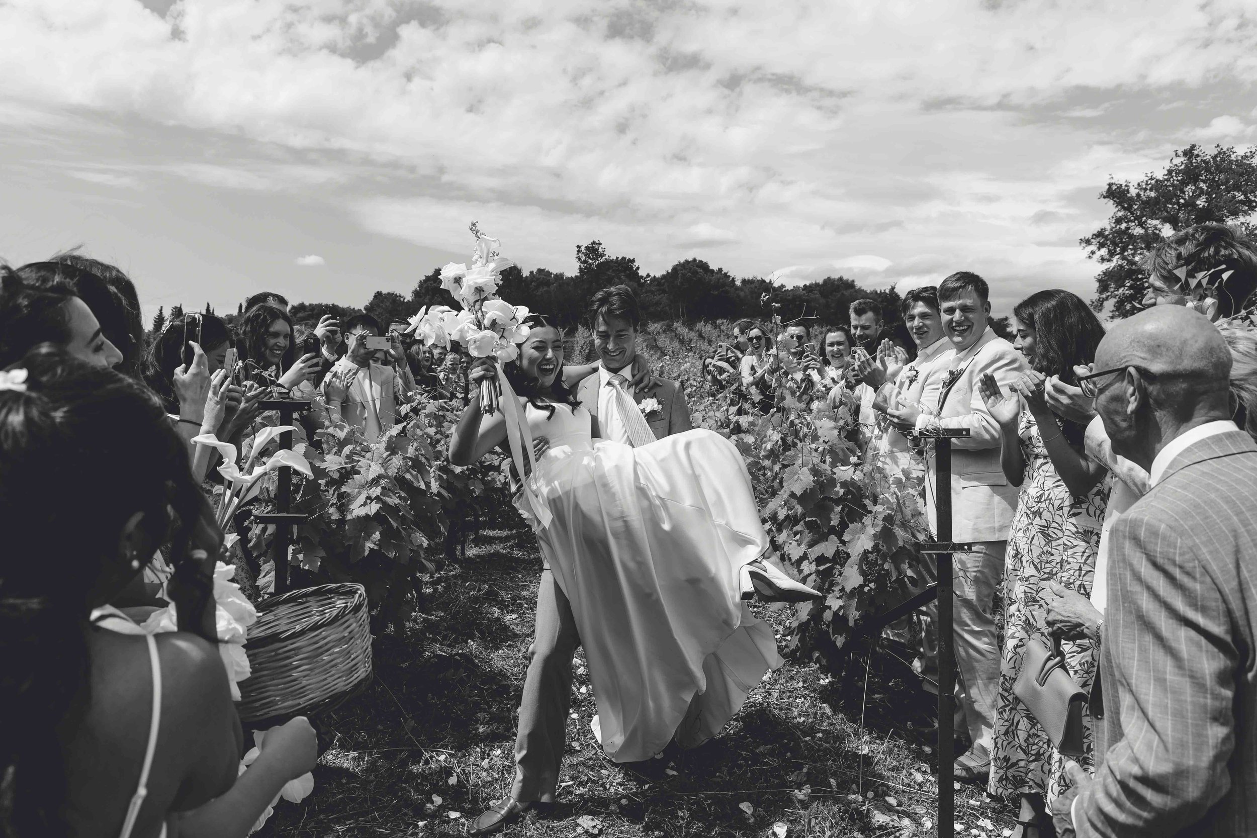 A wedding celebration outdoors with a bride being carried by the groom through a vineyard as friends and family cheer and take photos.