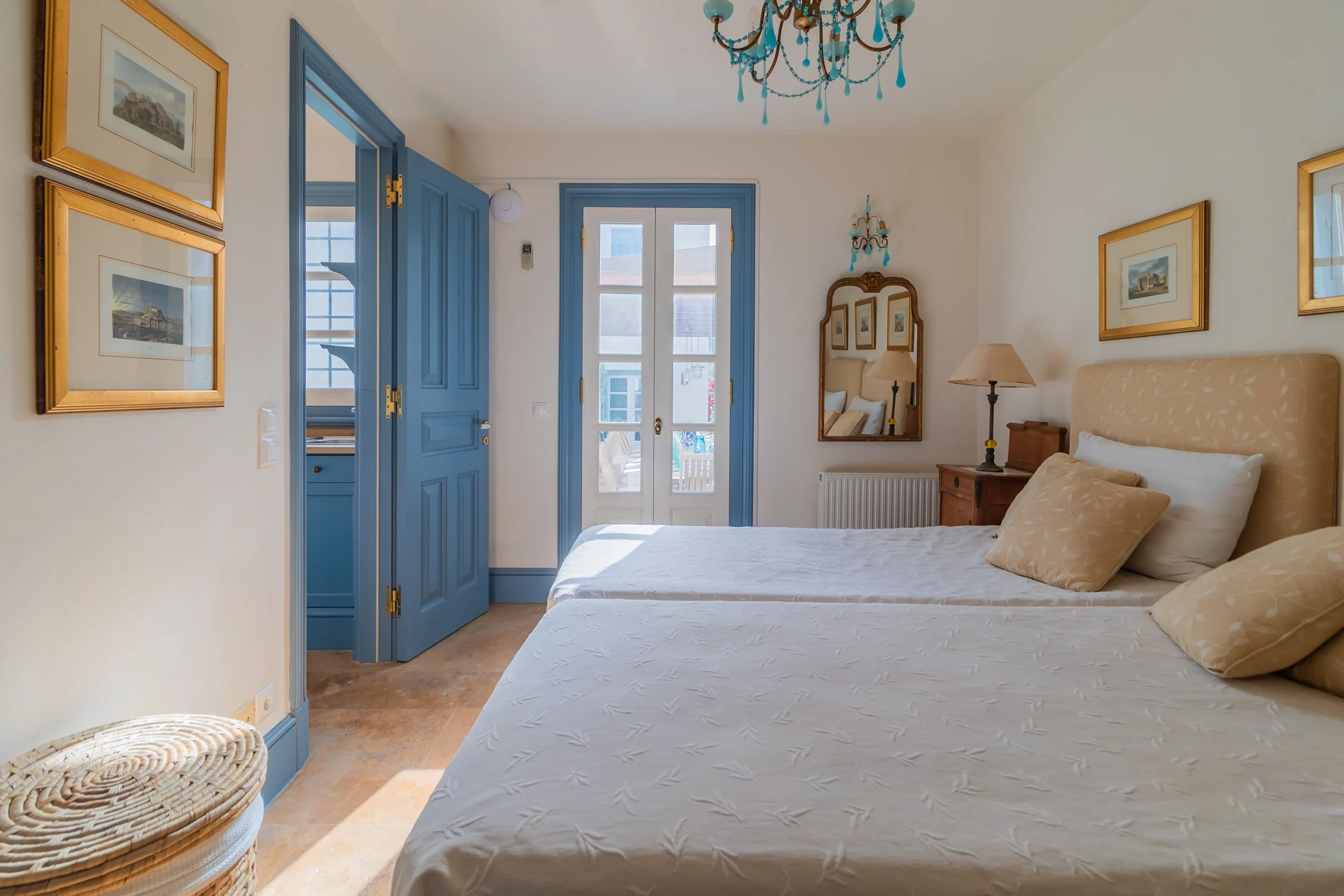 Cozy bedroom with two beds, beige headboards, cream bedding, and matching pillows. Blue-trimmed French doors lead outside, and a wooden bedside table with a lamp is beside the bed. Artworks hang on the cream walls, and a decorative chandelier hangs f