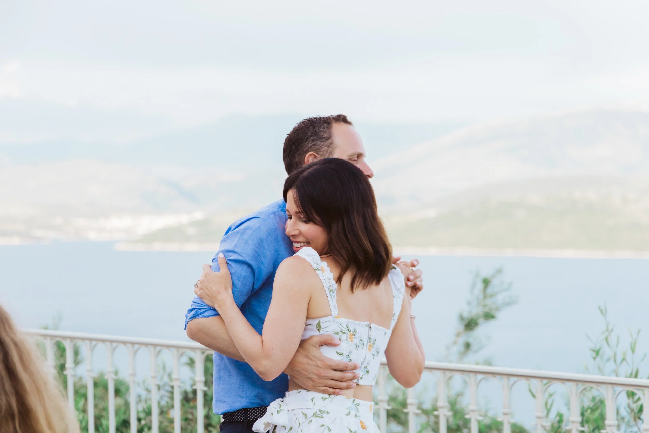 A couple embracing outdoors near a body of water, with a railing and distant mountains in the background.