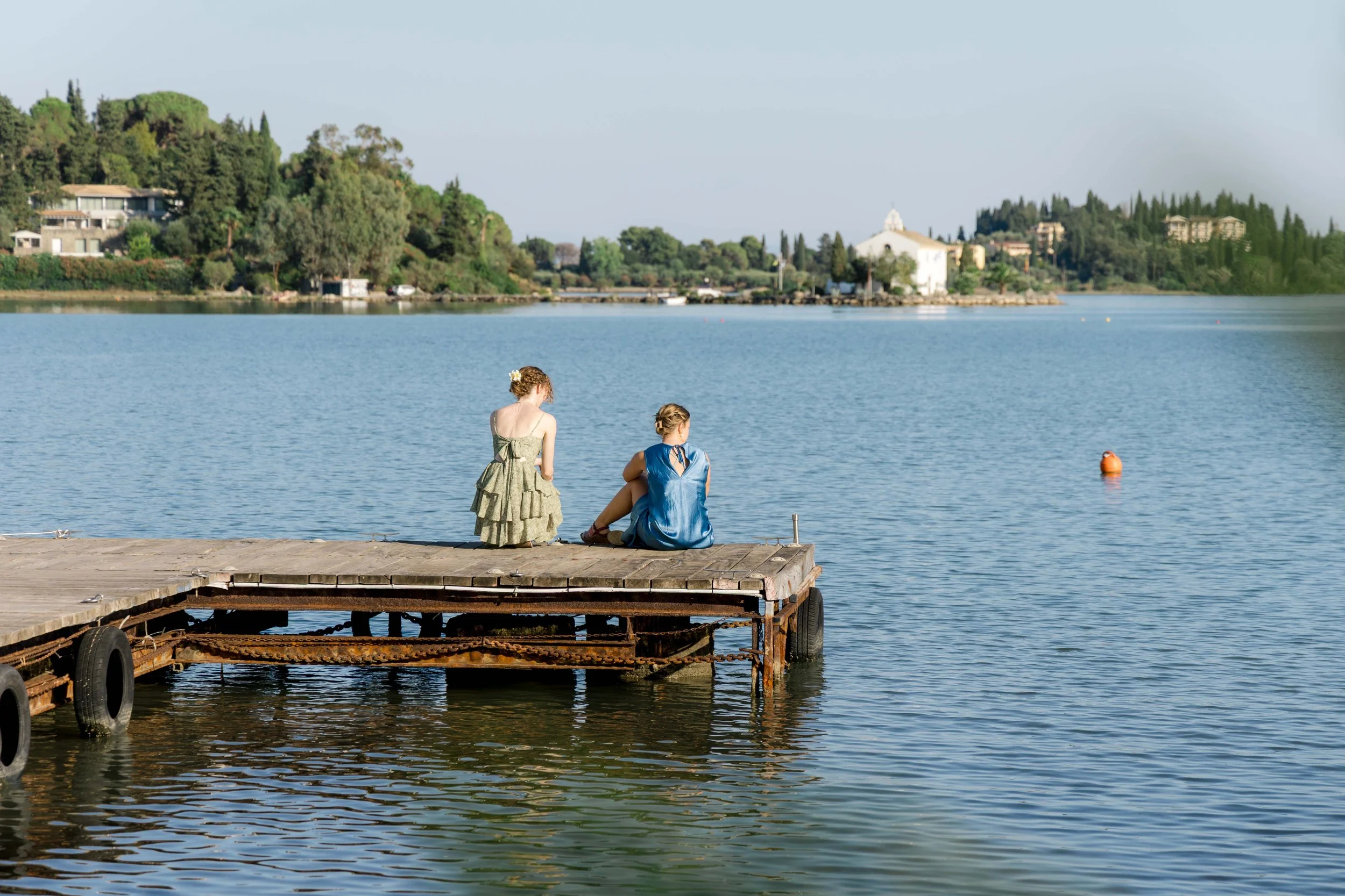 Two women sitting on a wooden dock by a calm lake, with a scenic hillside and buildings in the background on a sunny day.