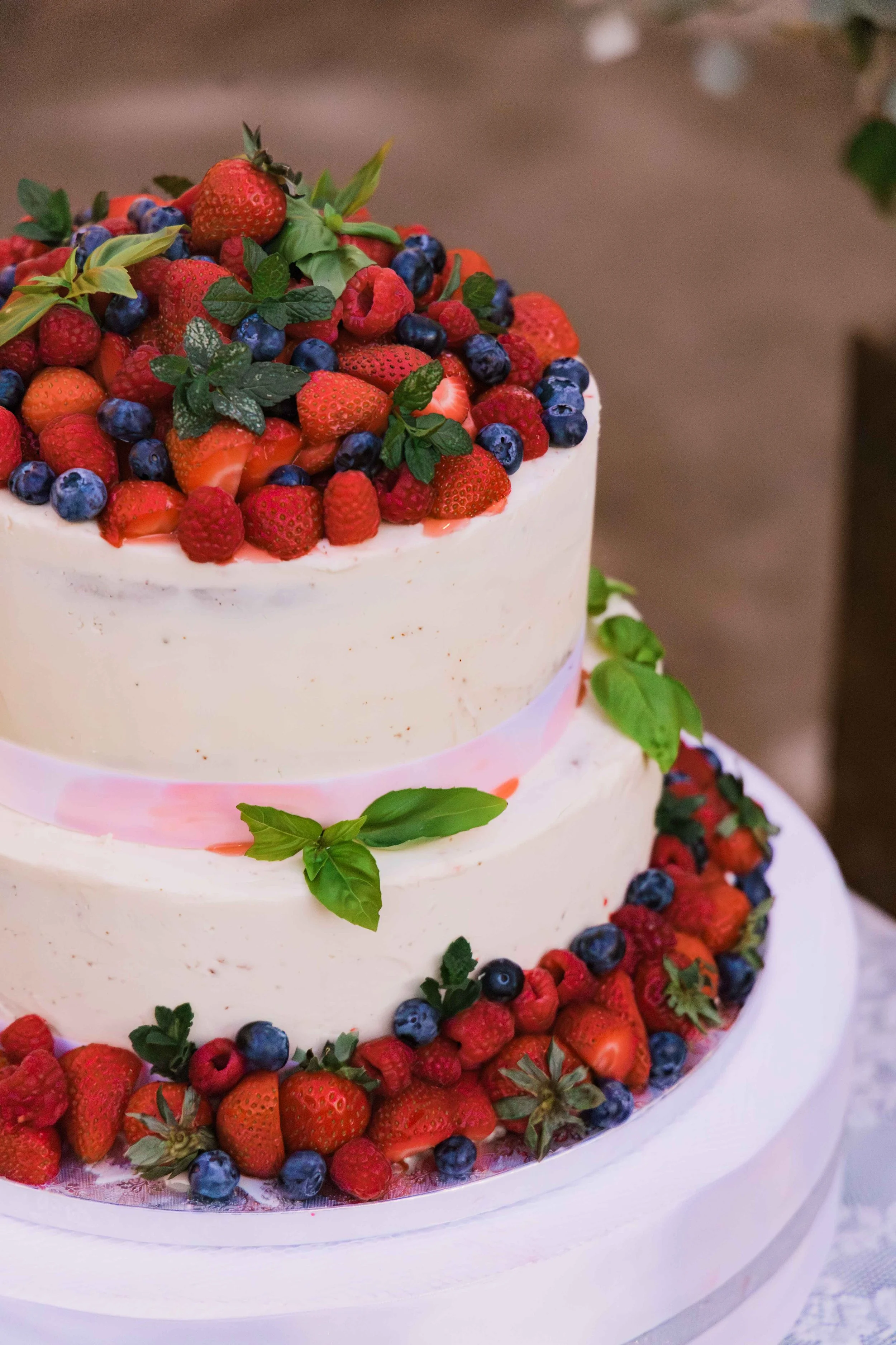 A two-tiered white wedding cake decorated with an assortment of fresh berries including strawberries, blueberries, raspberries, and blackberries, with a garnish of green leaves on top and around the base.