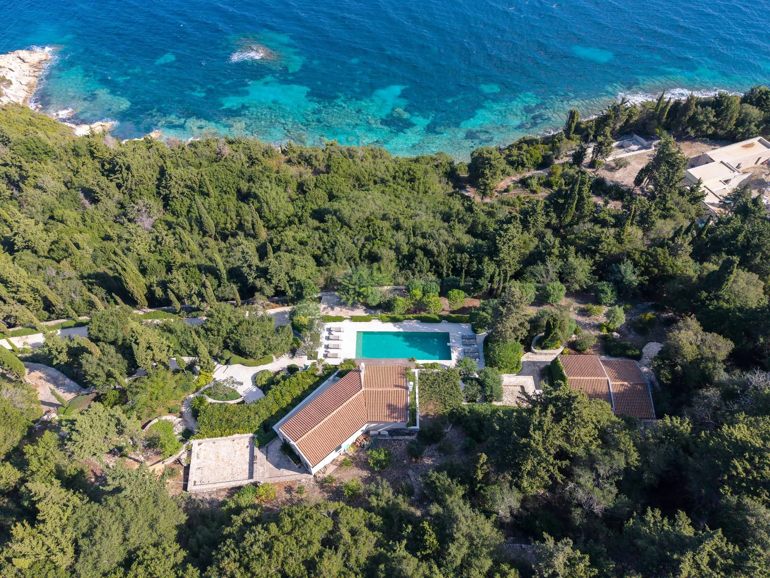 Aerial view of a house with a pool surrounded by lush green trees near the coastline with clear blue water.
