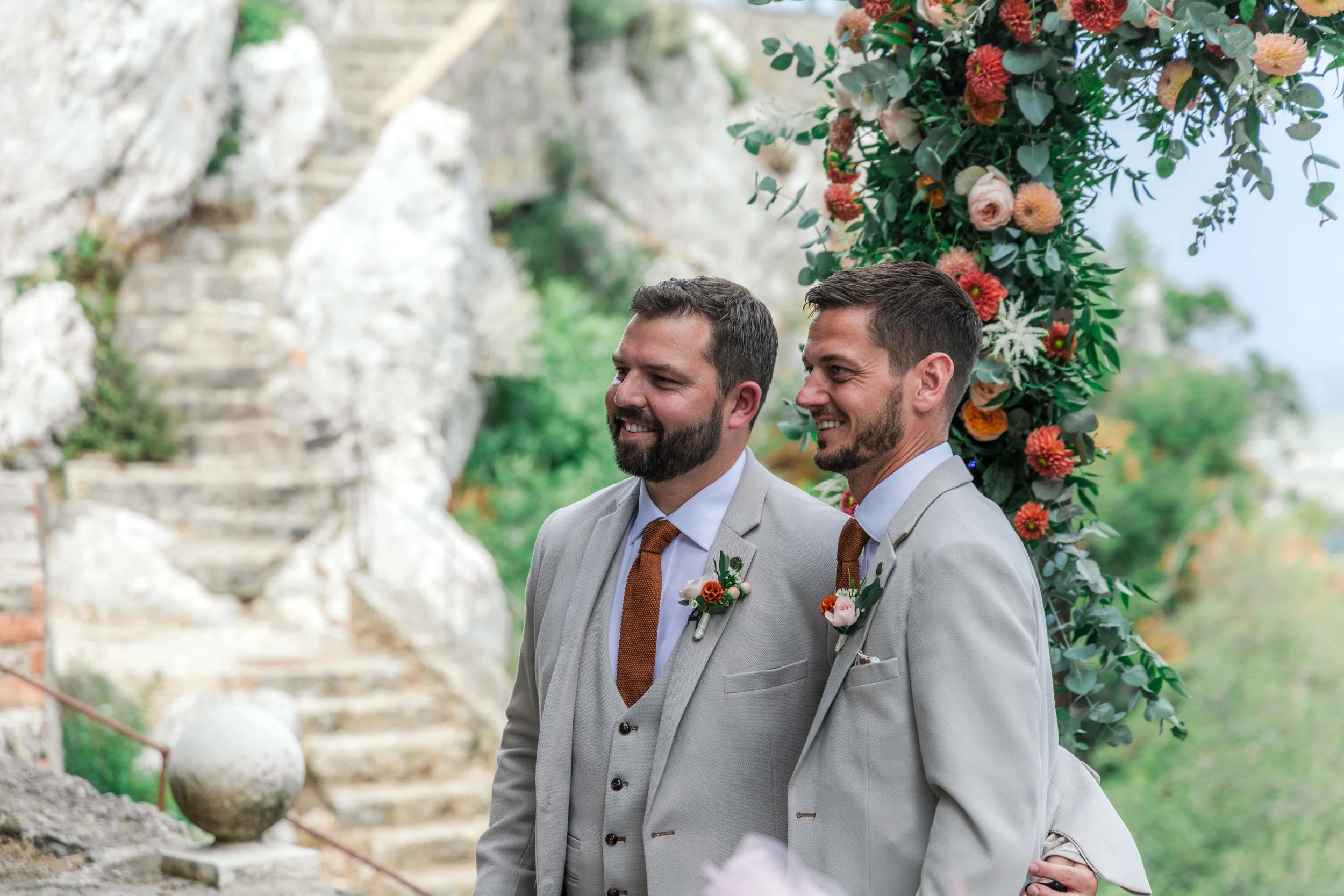 Two men in light-colored suits with boutonnières standing outdoors near a floral arch, smiling during a wedding ceremony.