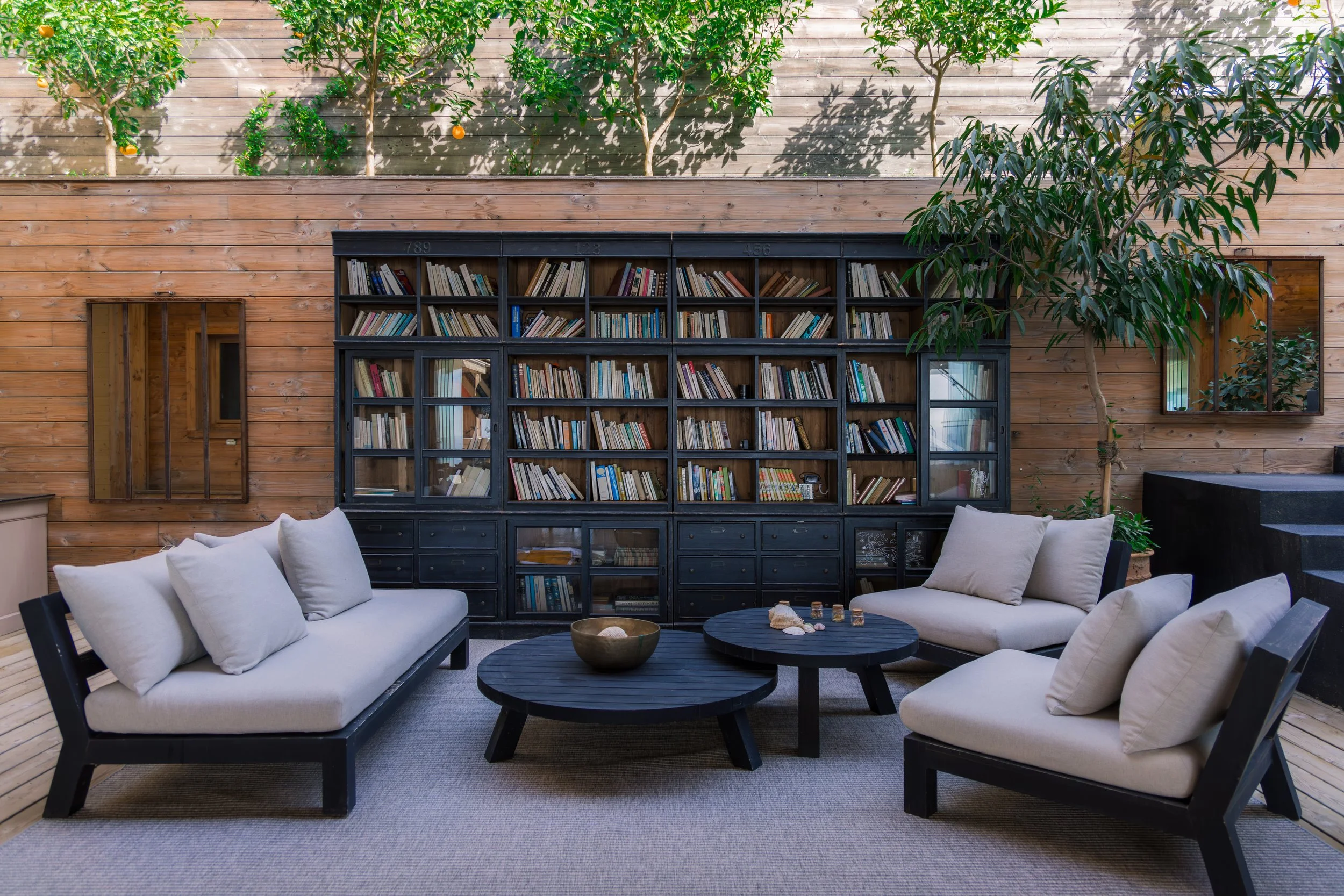 Indoor lounge area with black wooden furniture, beige cushions, a dark round coffee table with seashells, and a large black bookshelf filled with books. The background features a wooden wall with small trees and plants.