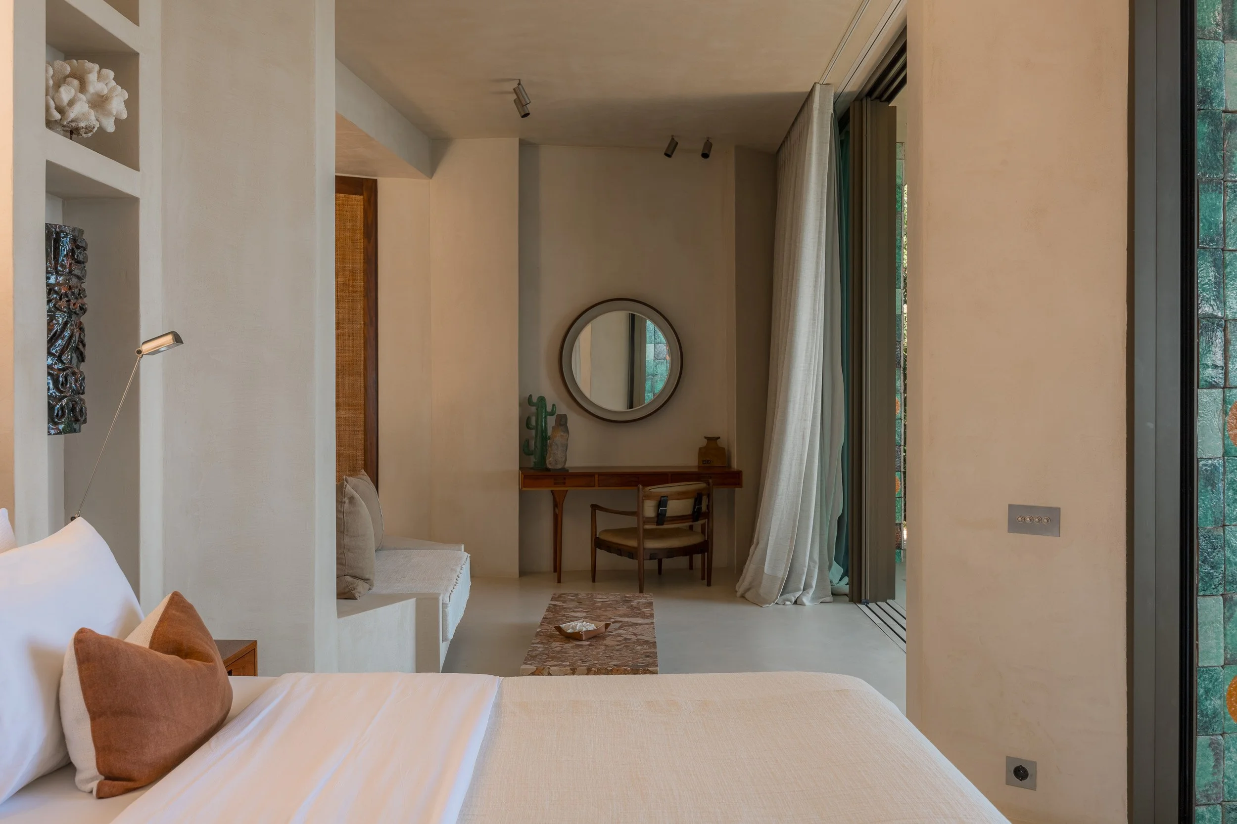Modern bedroom with a bed, pillows, a small bench, a wooden desk with a mirror and decorative vases, and a large window with curtains, natural light, and green foliage outside.
