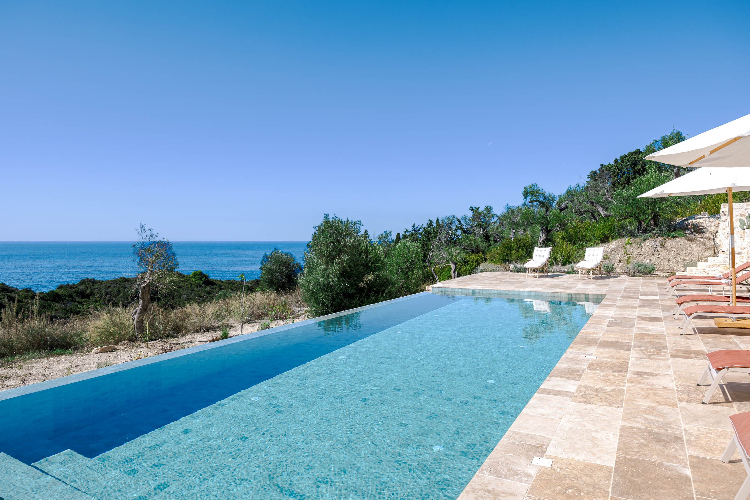 Infinity pool with ocean view, surrounded by a stone-tiled deck, a few lounge chairs with umbrellas, and green trees in the background under a clear blue sky.