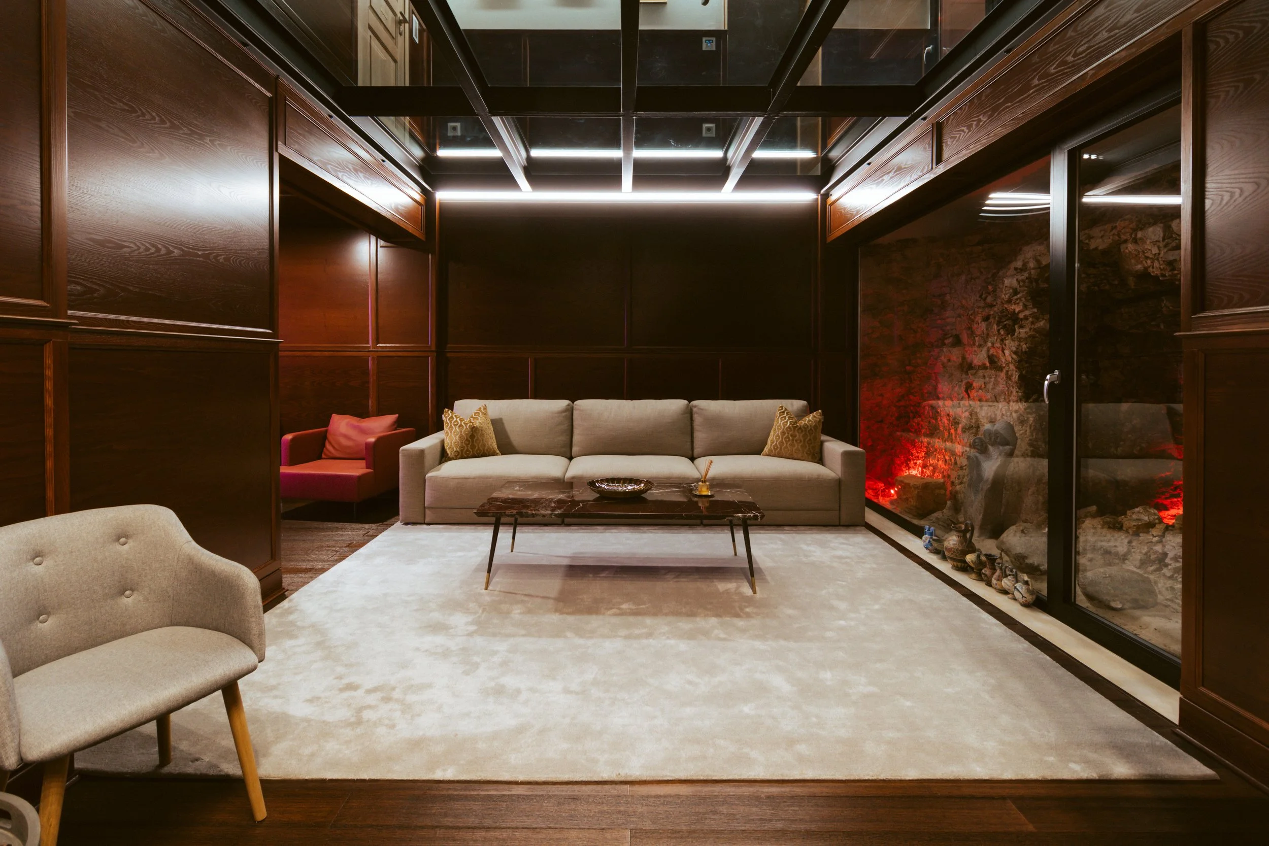 A modern living room with dark wood paneled walls, a beige sofa with decorative pillows, a small marble coffee table, a beige armchair, and a glass wall revealing a rock feature with red lighting.